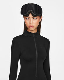 A woman wearing the Rudsak ZADIE - 8524609 Black long sleeve bodysuit and large black ski goggles on her head looks to the side against a plain white background.