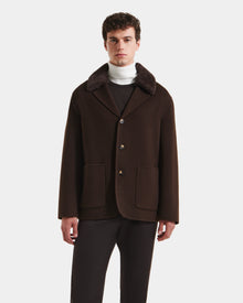 A man with short curly hair wears the Rudsak AIDEN - 6125558 Espresso wool-blend coat, featuring large buttons and front pockets, styled with dark pants and a white turtleneck against a white background.