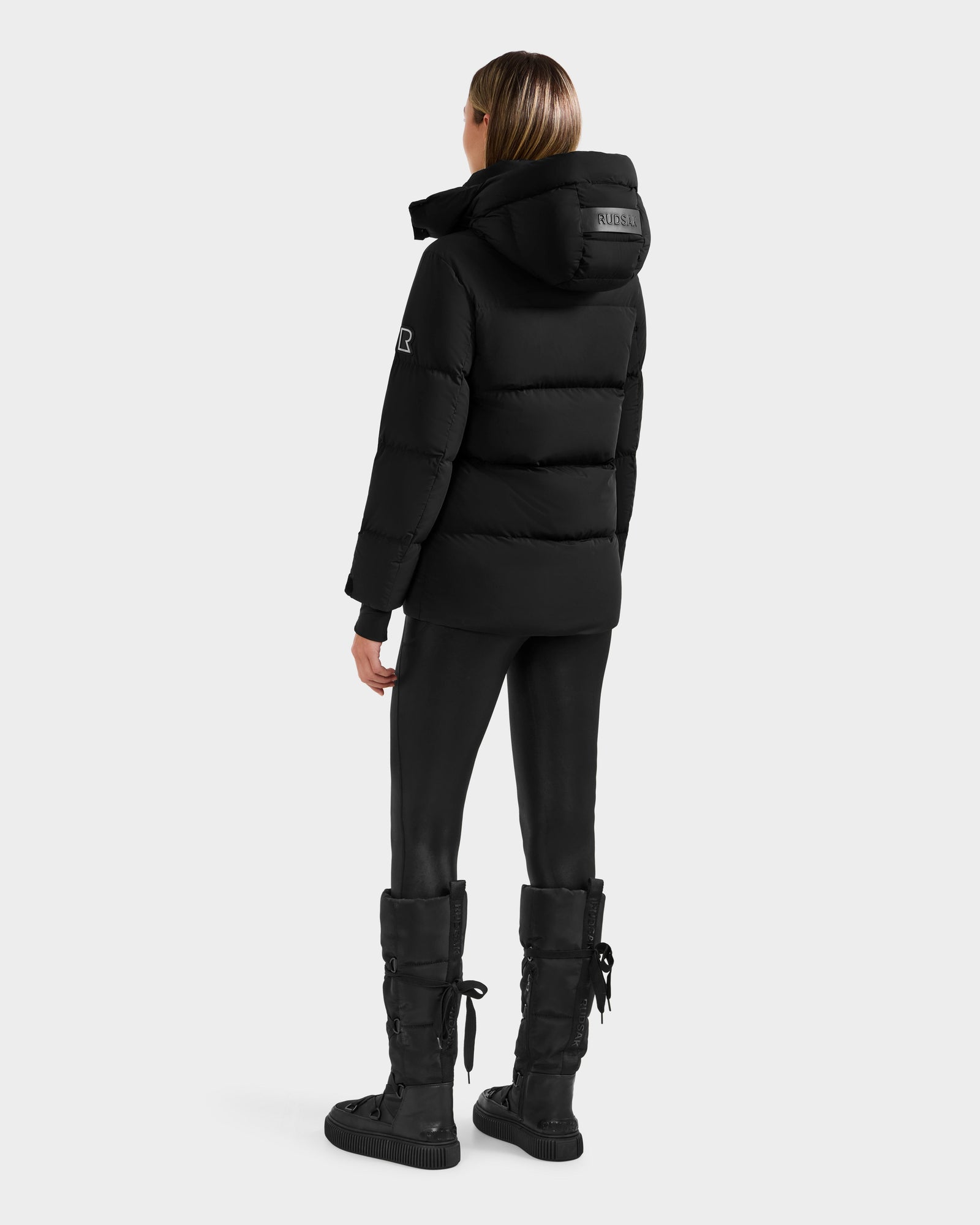 A person stands facing away in the Rudsak ALESSANDRA S-M - L125551 Black cropped puffer jacket with a hood, paired with black leggings and lace-up boots—a sleek, stylish cold-weather essential against a plain white backdrop.