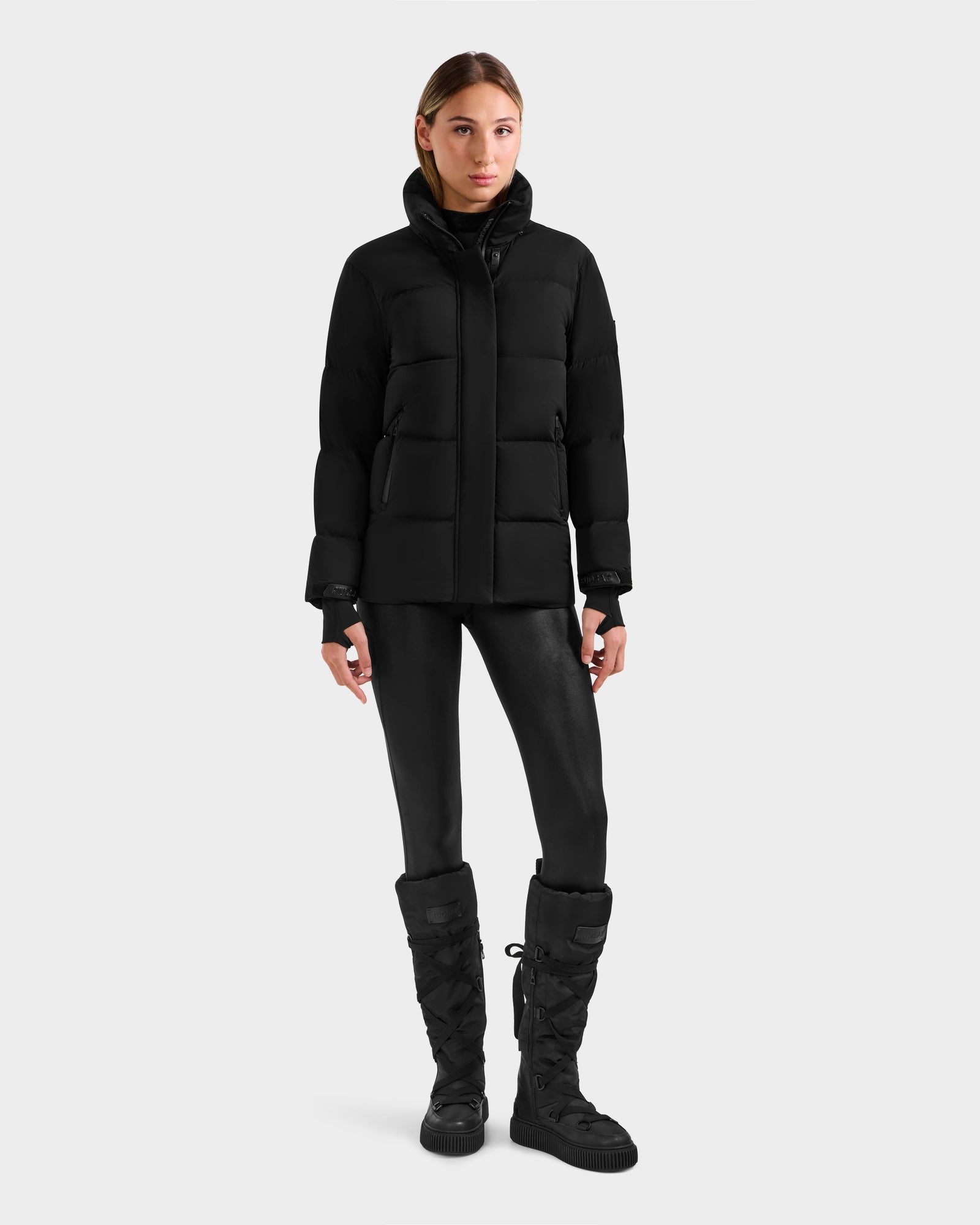A woman stands against a white background, wearing the Rudsak ALESSANDRA S-M - L125551 Black down puffer jacket with shiny leggings and black boots. Facing forward with arms relaxed, she showcases this cold-weather essential.