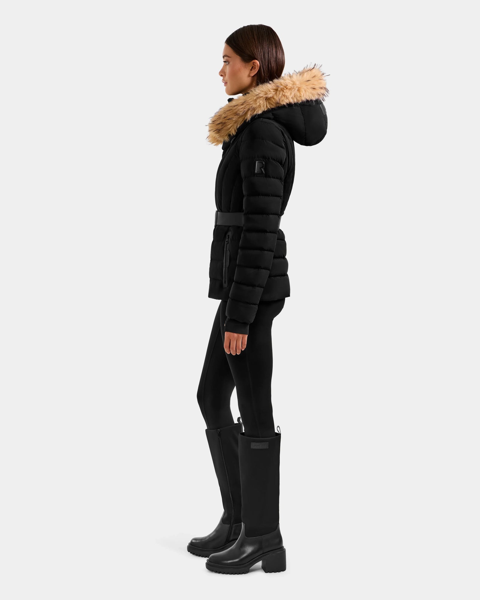 A woman in profile wears the Rudsak ALISA F - L125527 Black quilted puffer jacket with faux-fur trim hood, styled with black leggings and knee-high boots, standing against a plain light gray background.