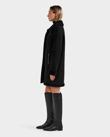 A woman stands in profile wearing the Rudsak ANGILICA - L125600 Black faux suede knee-length coat with faux shearling collar and cuffs, paired with tall black boots against a plain, light background.