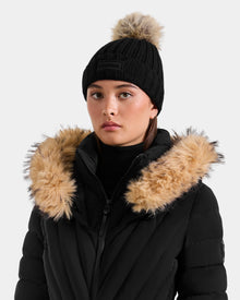 A person wears Rudsak’s ANISA - L423500 Black/Natural knit pom pom hat and a black puffer jacket with faux fur trim, standing against a plain light background—an essential winter look.
