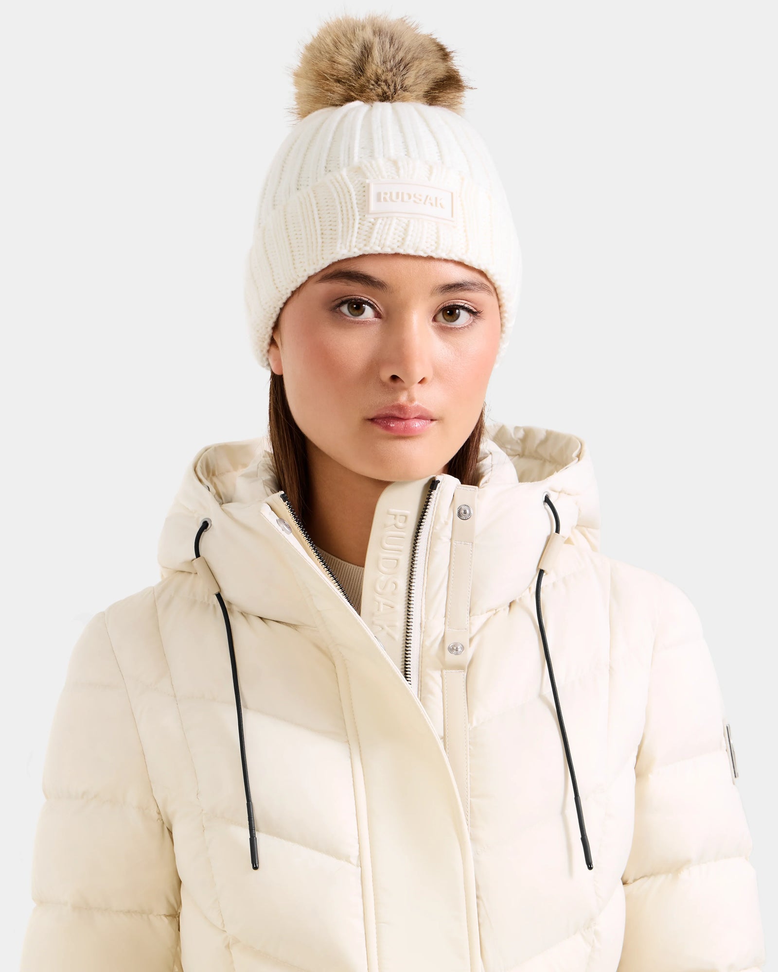 A woman models Rudsak’s ANISA - L423500 Optic White knit beanie with faux fur pom-pom and matching puffer jacket, both labeled Rudsak, against a light gray background—essential pieces for your winter wardrobe.