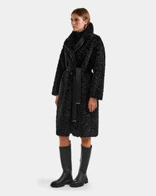 A person models the Rudsak ANNABELLE FSU - L125512 Black faux suede trench coat, featuring a wide collar and black belt, paired with black knee-high boots against a plain light background.