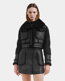 A woman models the Rudsak ARIELLA - 8125505 Black jacket with shearling trim and a matching black leather skirt, standing against a white background to showcase chic cold-weather style.