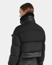 A person with straight brown hair models the Rudsak ARIELLA - 8125505 Black women’s cropped shearling puffer, featuring a high collar and leather accents, against a plain light background—perfect for cold-weather styling.