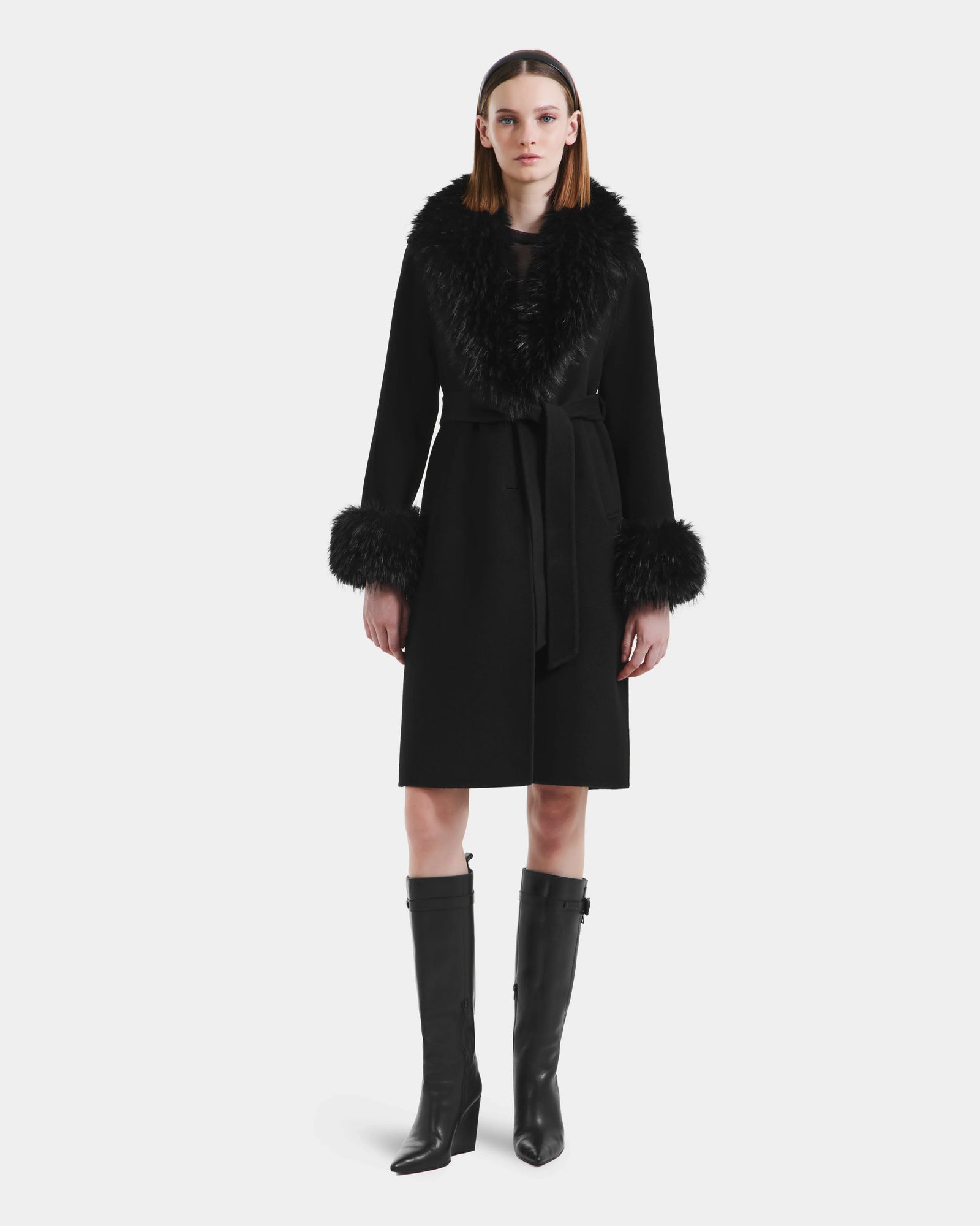 A woman stands facing forward in the Rudsak ARTIS - L125575 Black women’s wool coat with a belted waist, wide faux fur collar, fur-trimmed cuffs, and tall black heeled boots against a plain white background.