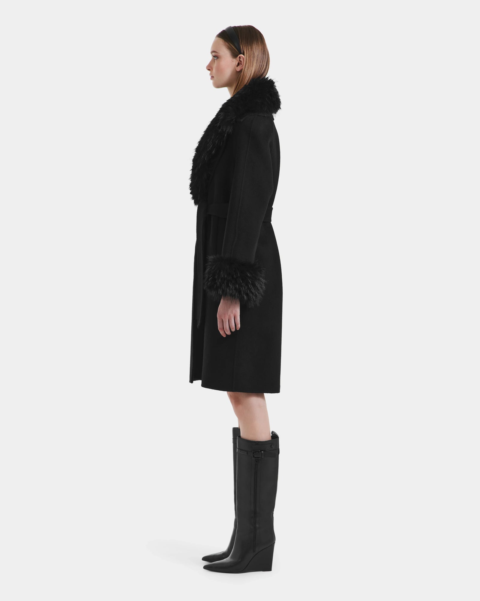 A woman stands in profile wearing the ARTIS - L125575 Black women’s wool coat by Rudsak, featuring a faux fur collar and cuffs, belted waist, black knee-high boots, and a black headband against a plain white background.