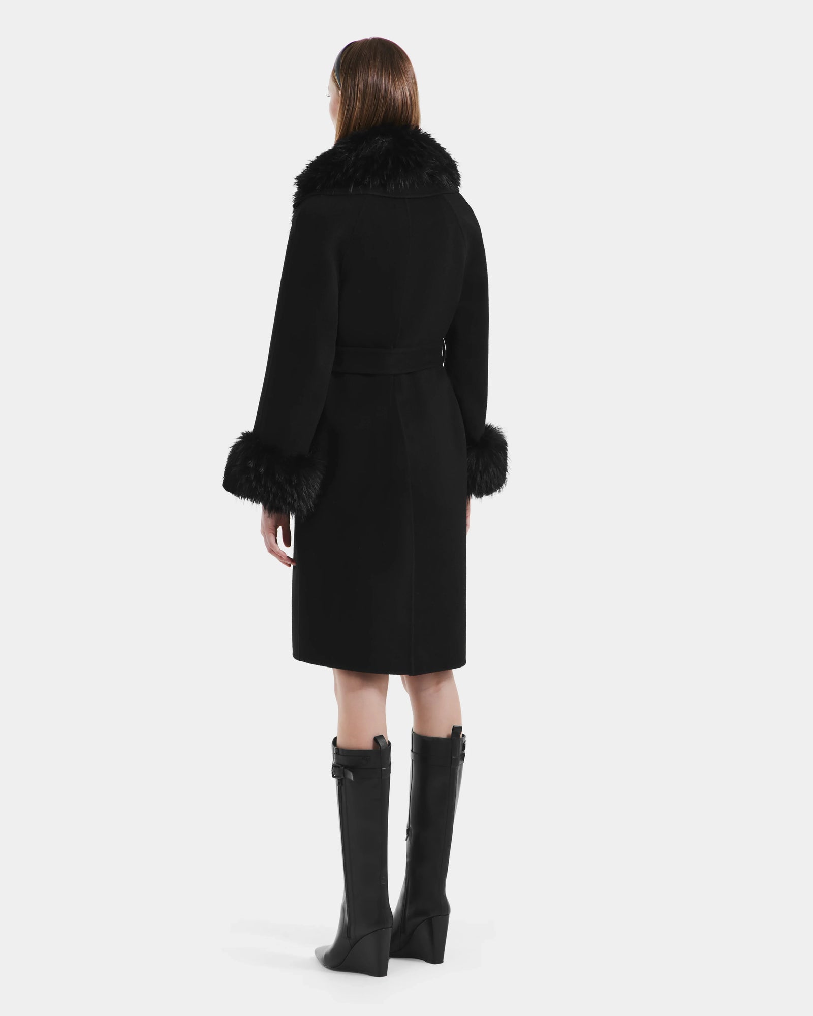 A woman with straight brown hair wears the Rudsak ARTIS - L125575 Black wool coat, featuring a belted waist and faux fur collar and cuffs, styled with knee-high black heeled boots against a plain white background.