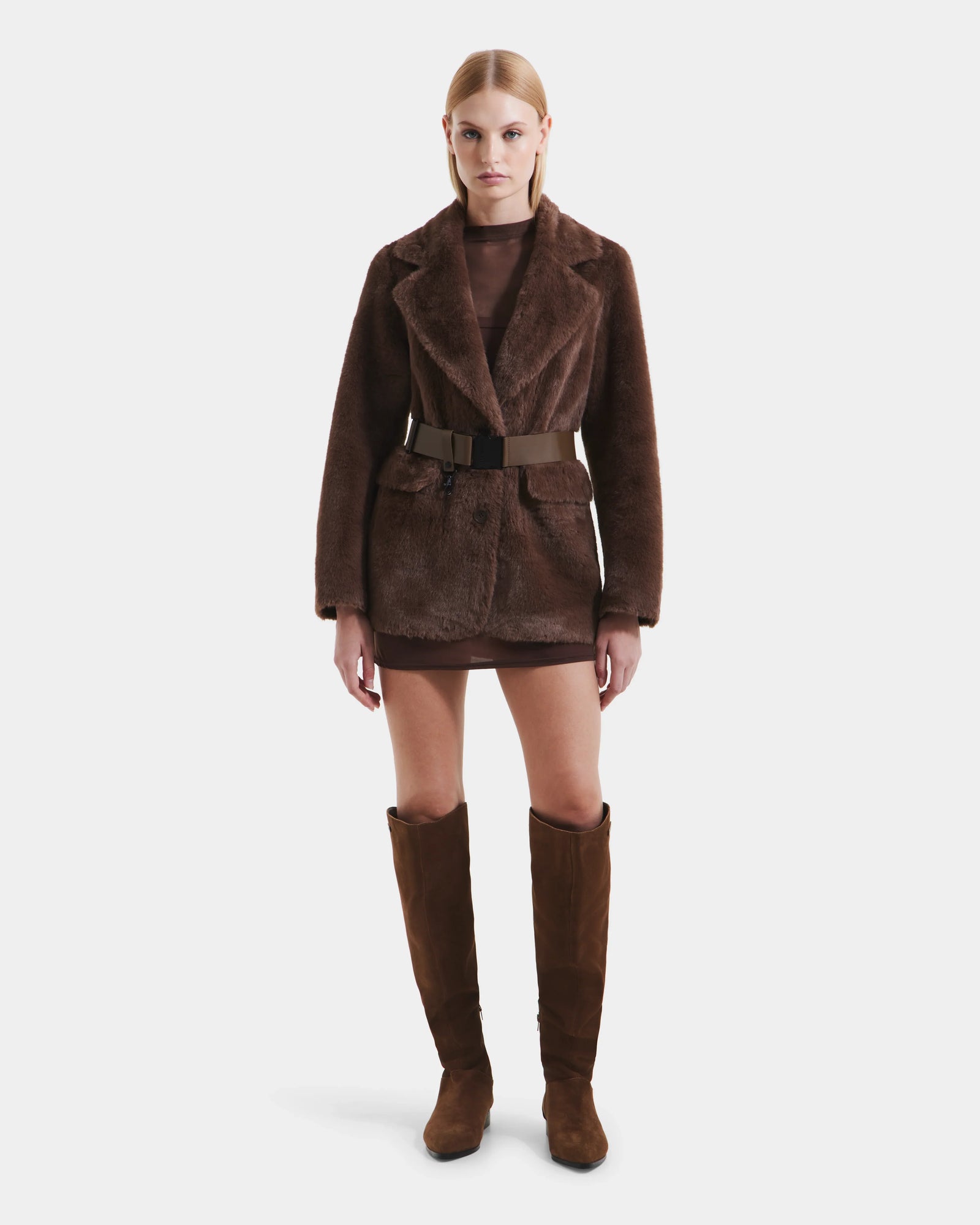 A woman faces forward in the Rudsak ASPEN F - 8125604 Mocha faux fur jacket with a wide belt, paired with a short brown dress and knee-high suede boots—an elevated cold-weather look—set against a plain white background.