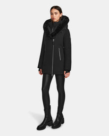 A woman stands against a plain white background wearing the Rudsak AURORE - L123561 Black/Black women's short fitted down parka with a fur-trimmed hood, paired with black leggings and black boots.
