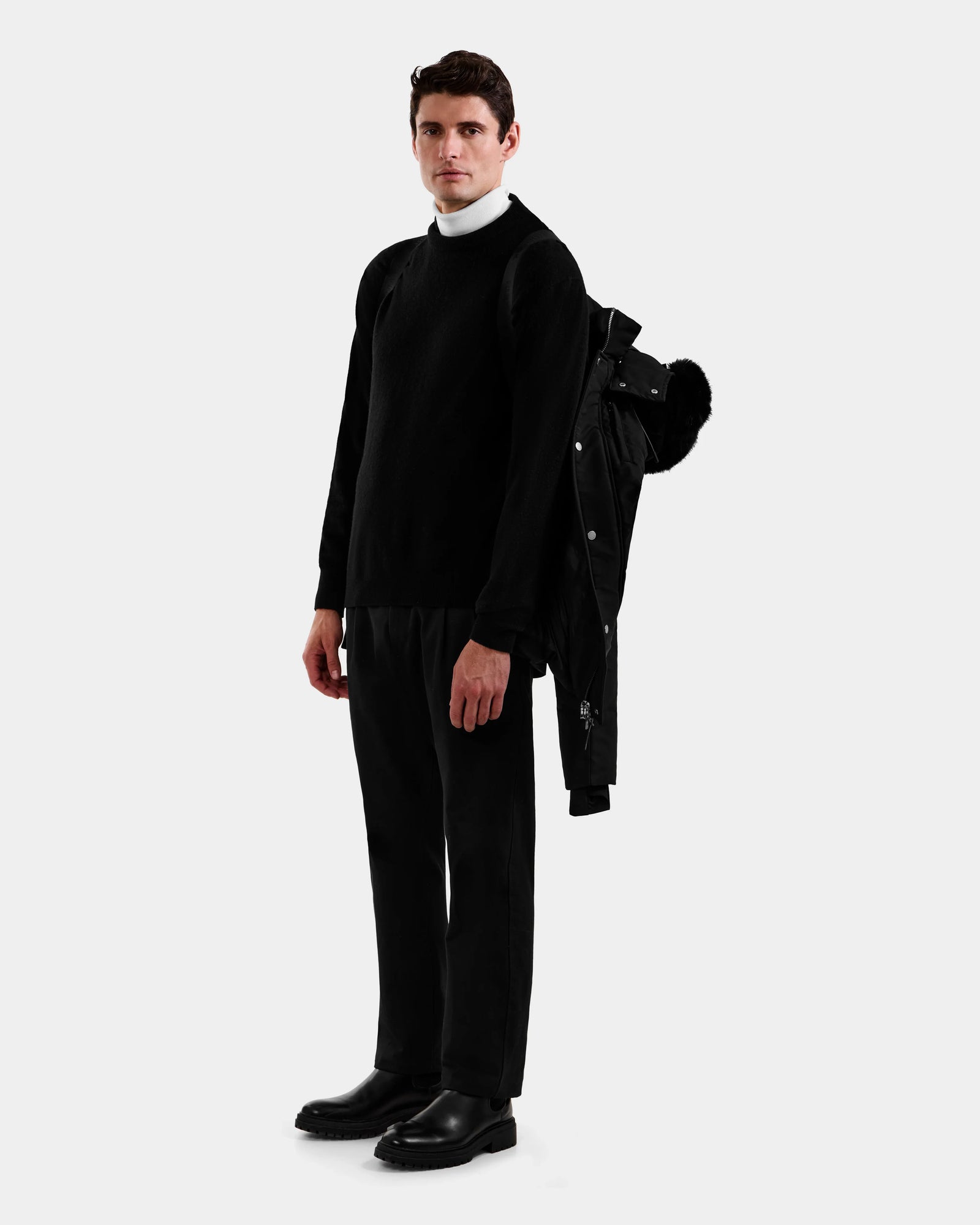 A man faces forward wearing a black sweater over a white turtleneck, black pants and shoes. The Rudsak BRADY - M125510 Black men’s quilted down parka with removable hood is draped over his left shoulder against a plain light gray background.