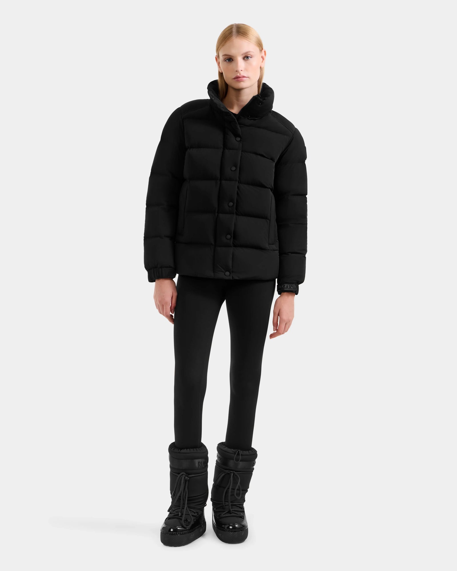 A woman with straight blond hair models the Rudsak BREE M - L125582 Black women’s down puffer jacket with a removable hood, styled with black leggings and boots against a plain light gray background—a cold-weather essential.