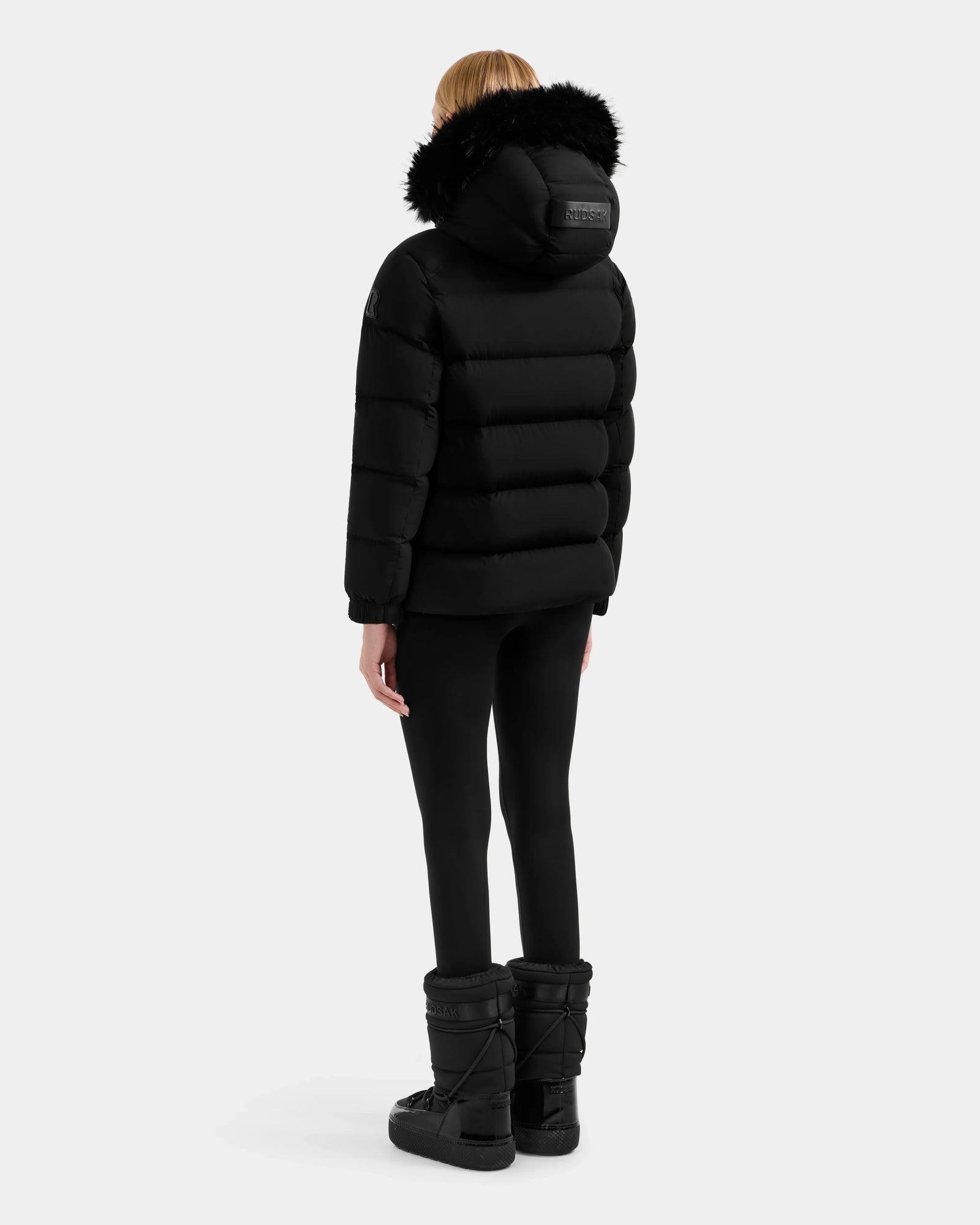 A person stands with their back turned, wearing the Rudsak BREE M - L125582 Black women’s down puffer jacket with a fur-trimmed removable hood, black leggings, and winter boots—an essential cold-weather look on a white background.