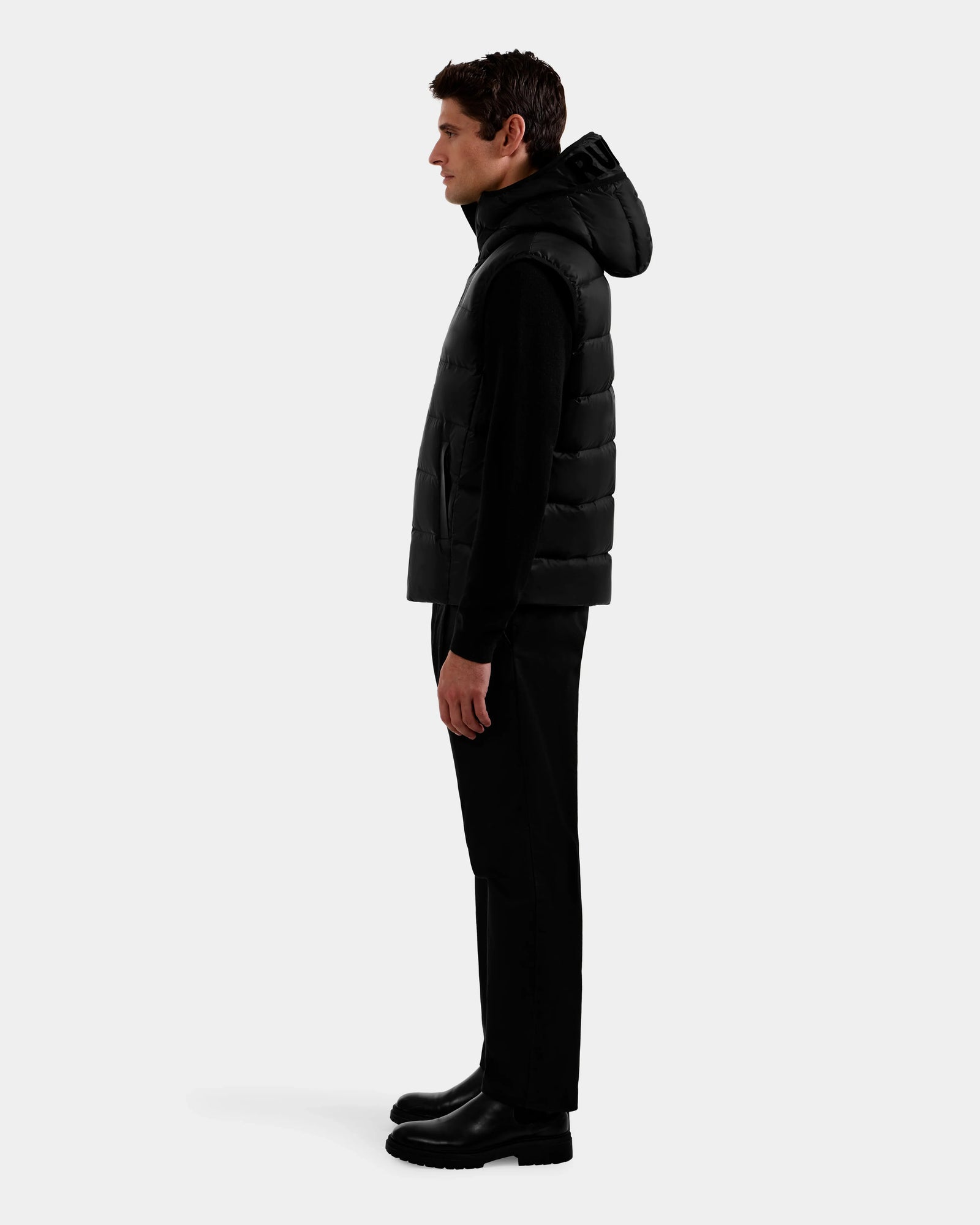 A man stands in profile facing left, wearing the Rudsak BROOKS - M125520 Black men’s down puffer vest with a recycled-nylon shell, paired with black pants and boots against a plain light gray background.