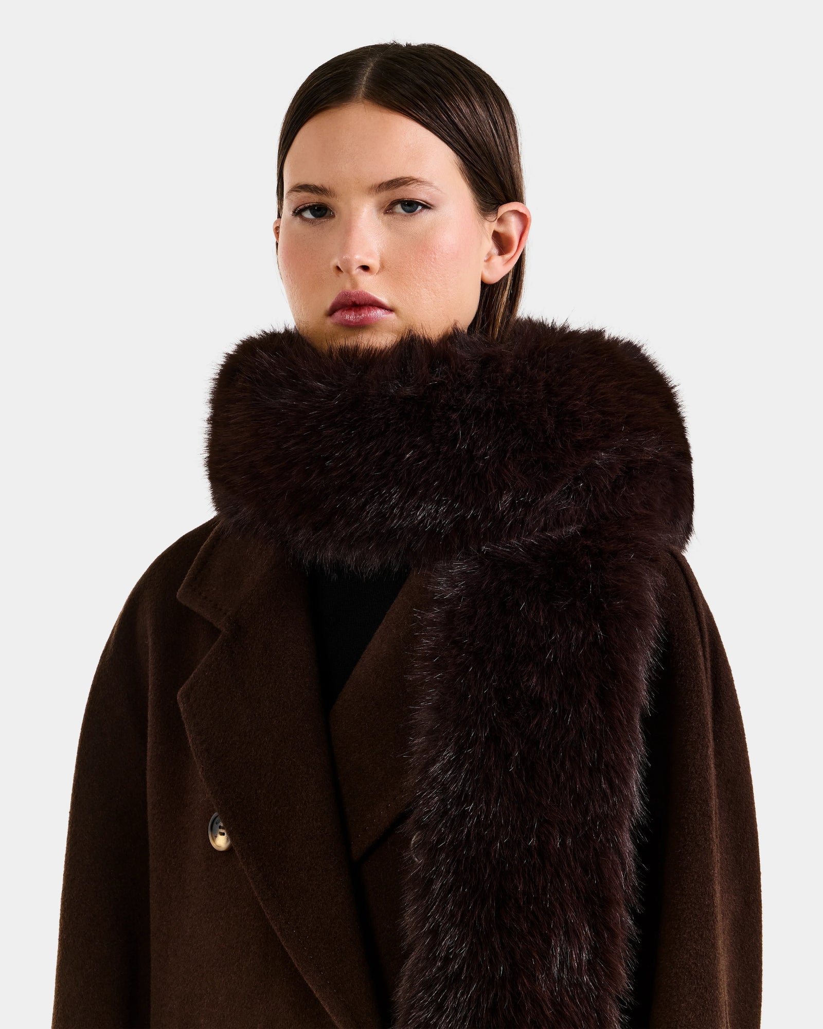 A woman with straight brown hair models the Rudsak CARMY - L425534 Espresso, a plush women’s faux-fur scarf, wrapped around her neck and paired with a dark brown coat against a plain light background.