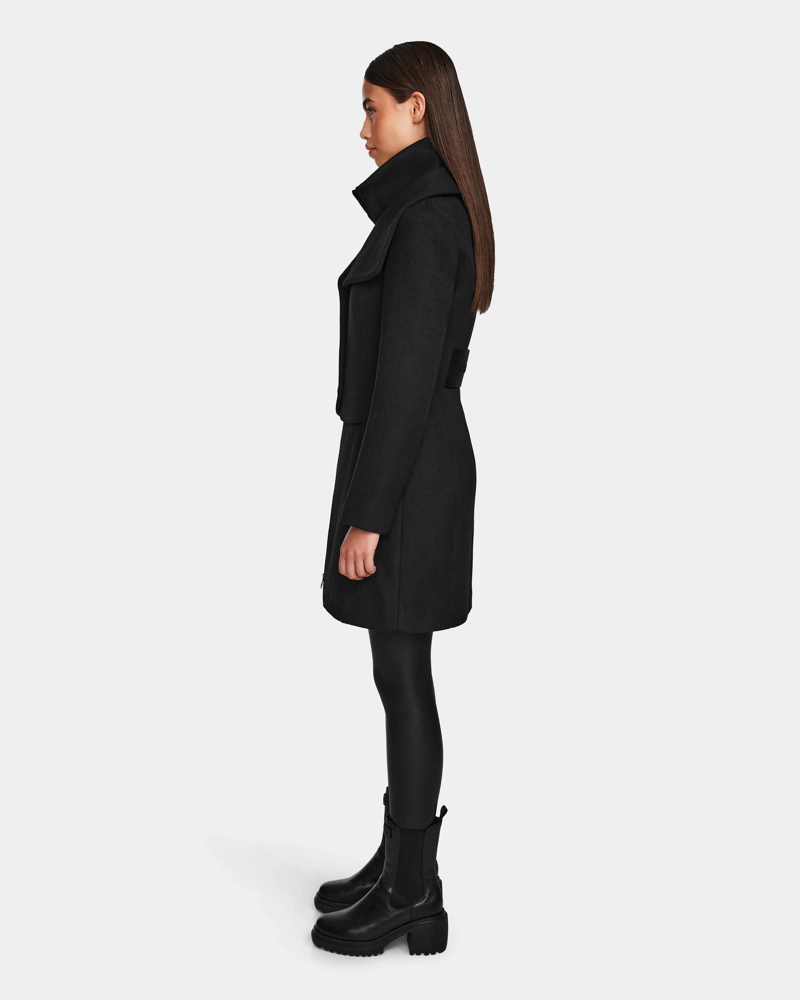 A woman stands in profile wearing the Rudsak CAROLINE - L124543 Black knee-length premium wool blend coat, paired with black leggings and chunky boots against a plain light background.