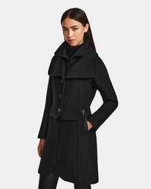 A woman with long dark hair pulled back and a neutral expression wears the Rudsak CAROLINE - L124543 Black, a chic knee-length premium wool-blend coat with large buttons, a wide collar, and black vegan leather-trimmed pockets.
