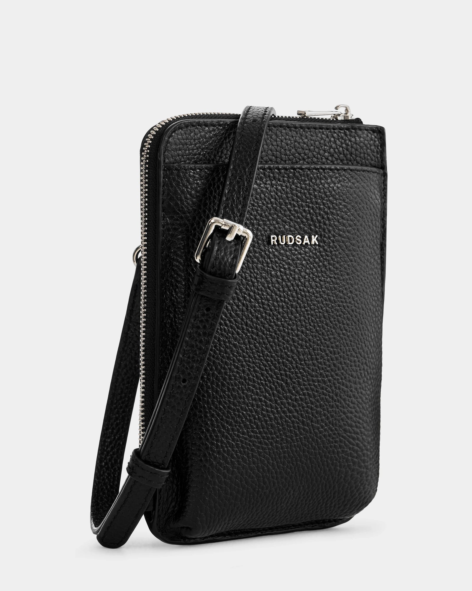 The Rudsak CELYN - L325514 Black is a women's black pebbled leather crossbody bag with a slim rectangular shape, silver buckle, zipper closure, adjustable strap, and Rudsak logo on the front.