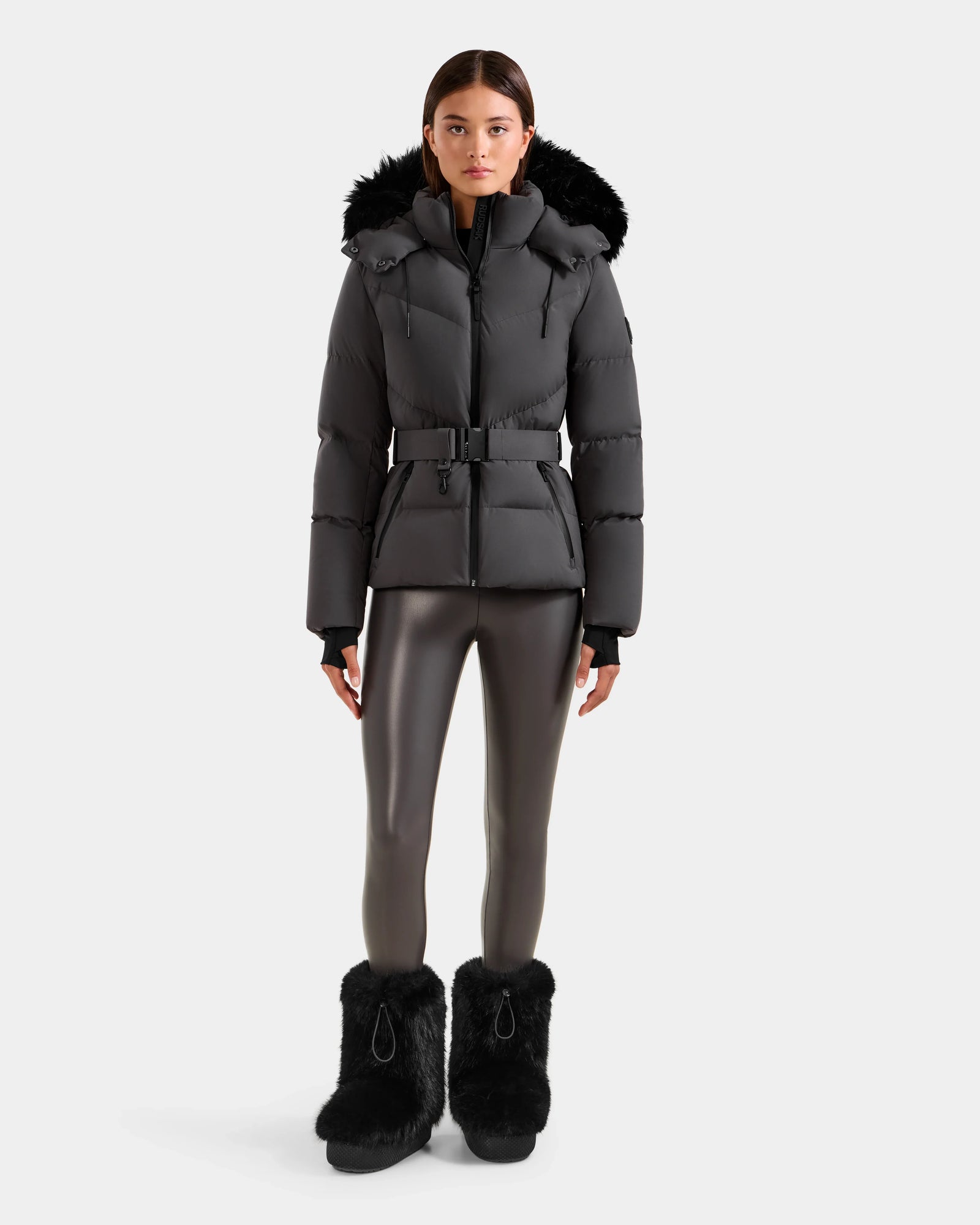A person stands facing forward against a plain white background wearing the Rudsak CHRISTELLE M - L125542 Grey weather-resistant quilted down puffer jacket with a fur-trimmed hood.