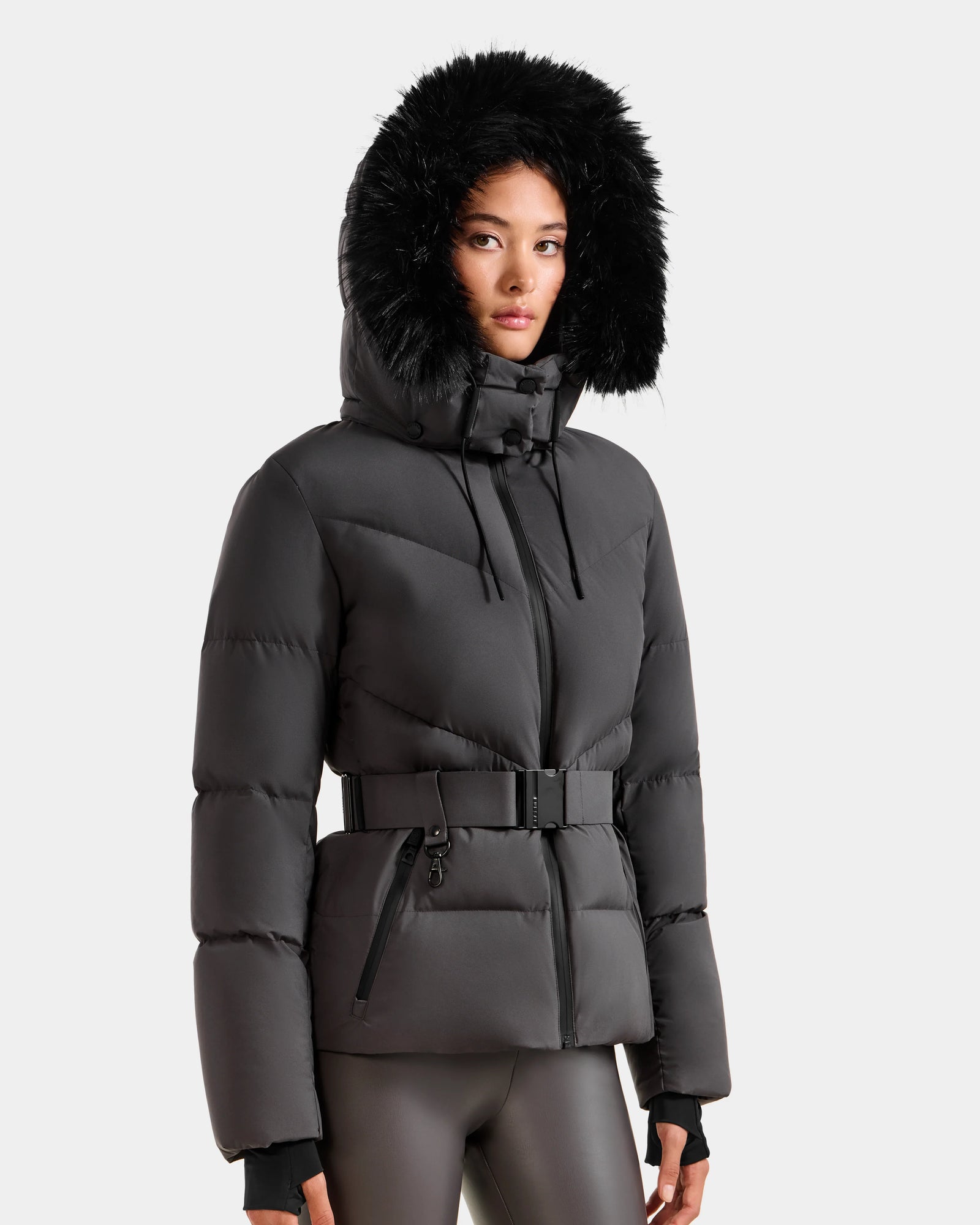 A woman wearing the Rudsak CHRISTELLE M - L125542 Grey quilted down puffer jacket with faux fur trim, front zipper pockets, and belted waist stands against a plain light gray background, paired with black leggings.