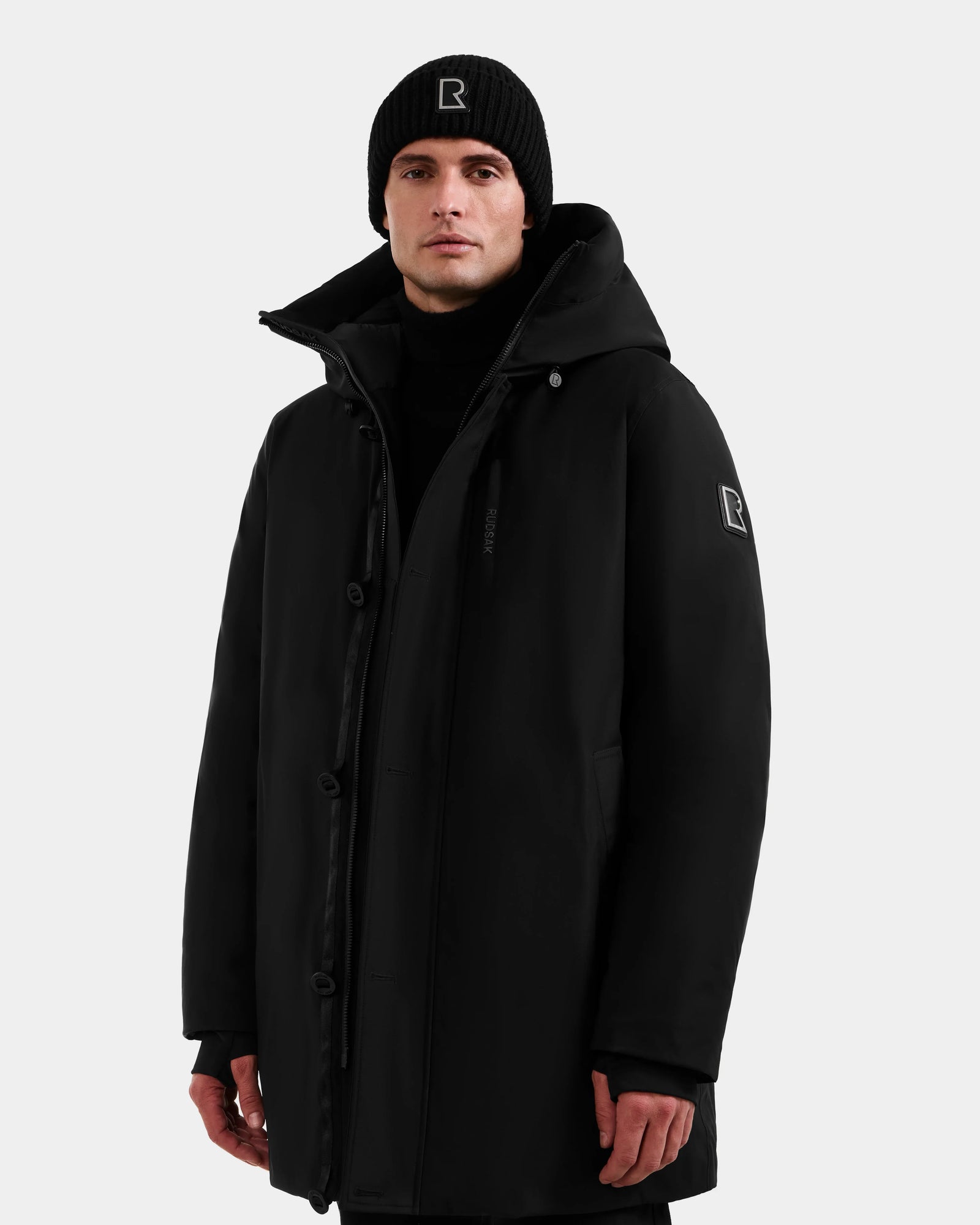 A man stands against a light gray background wearing Rudsak’s COREY - M125512 Black down parka and a black beanie with a white R logo, looking forward with a neutral expression.
