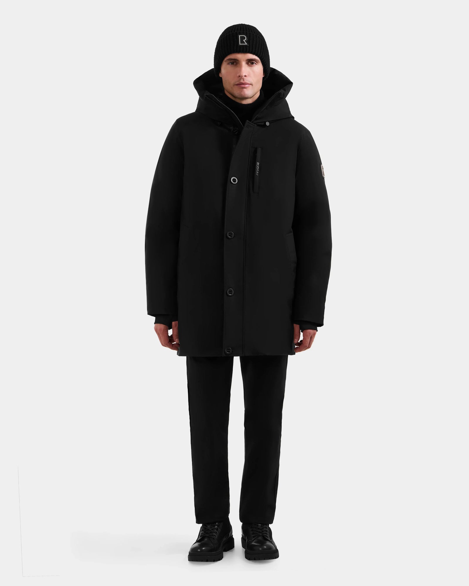 A person wearing the Rudsak COREY - M125512 Black men’s down parka, black pants, shoes, and beanie stands facing forward against a plain light gray background.