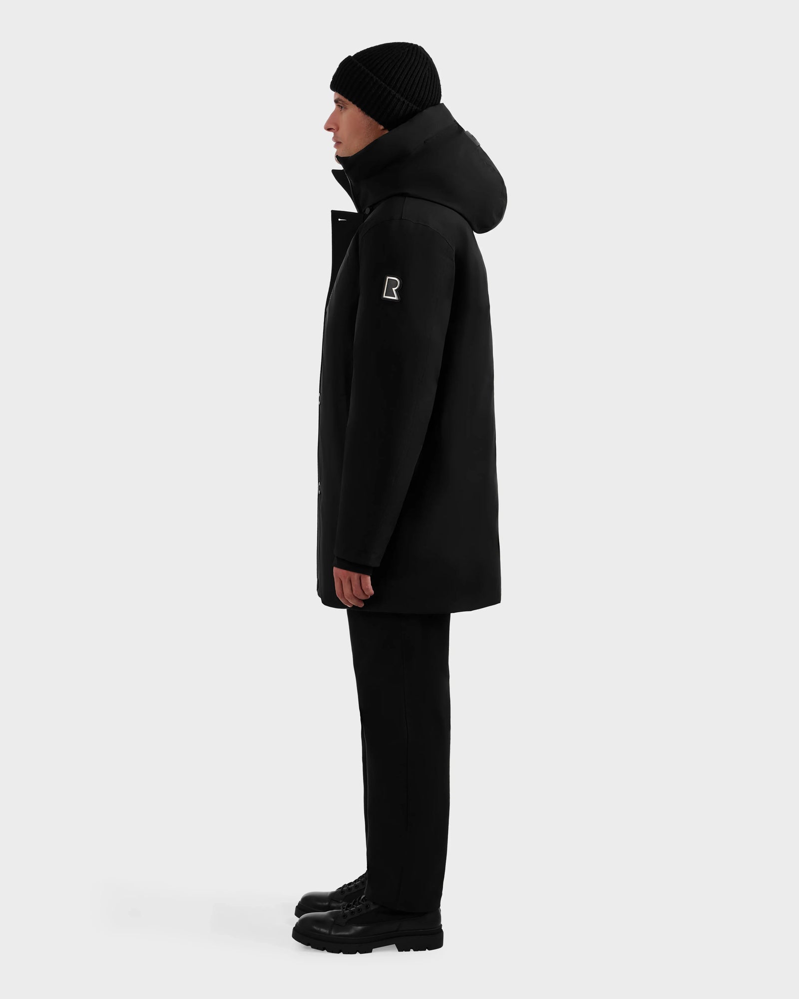 A person in profile wears the Rudsak COREY - M125512 Black men’s down parka with a logo patch on the sleeve, black beanie, pants, and boots, standing against a plain white background.