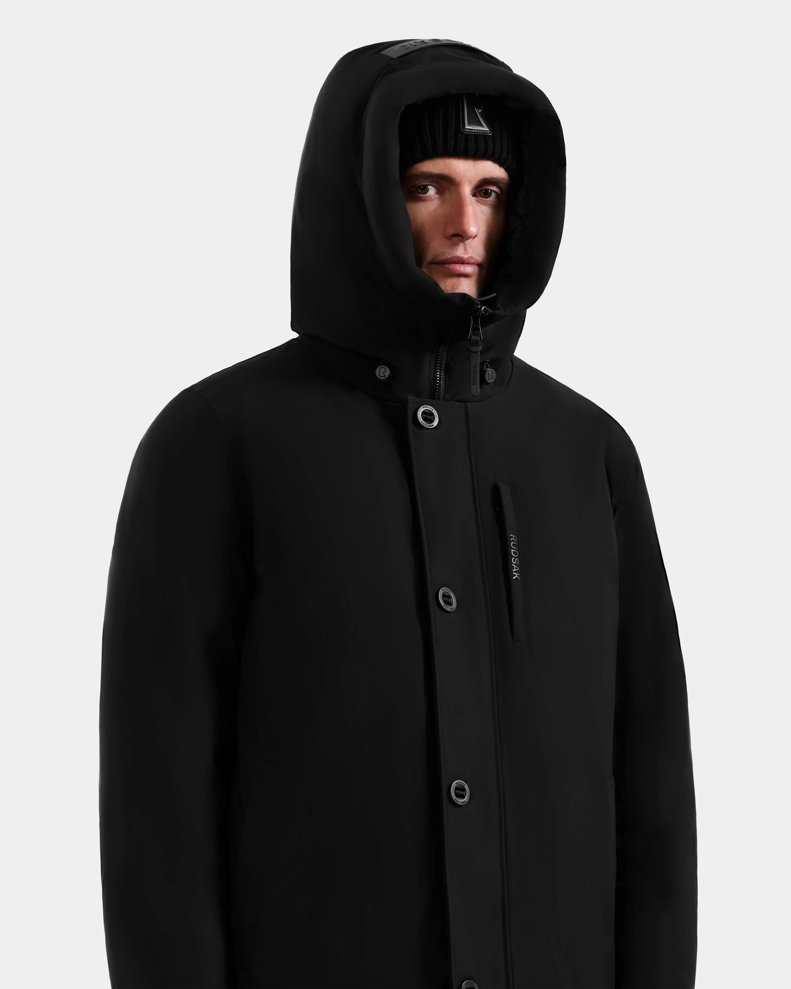A person wearing the Rudsak COREY - M125512 Black men’s down parka and a black beanie looks slightly to the side against a plain light background.