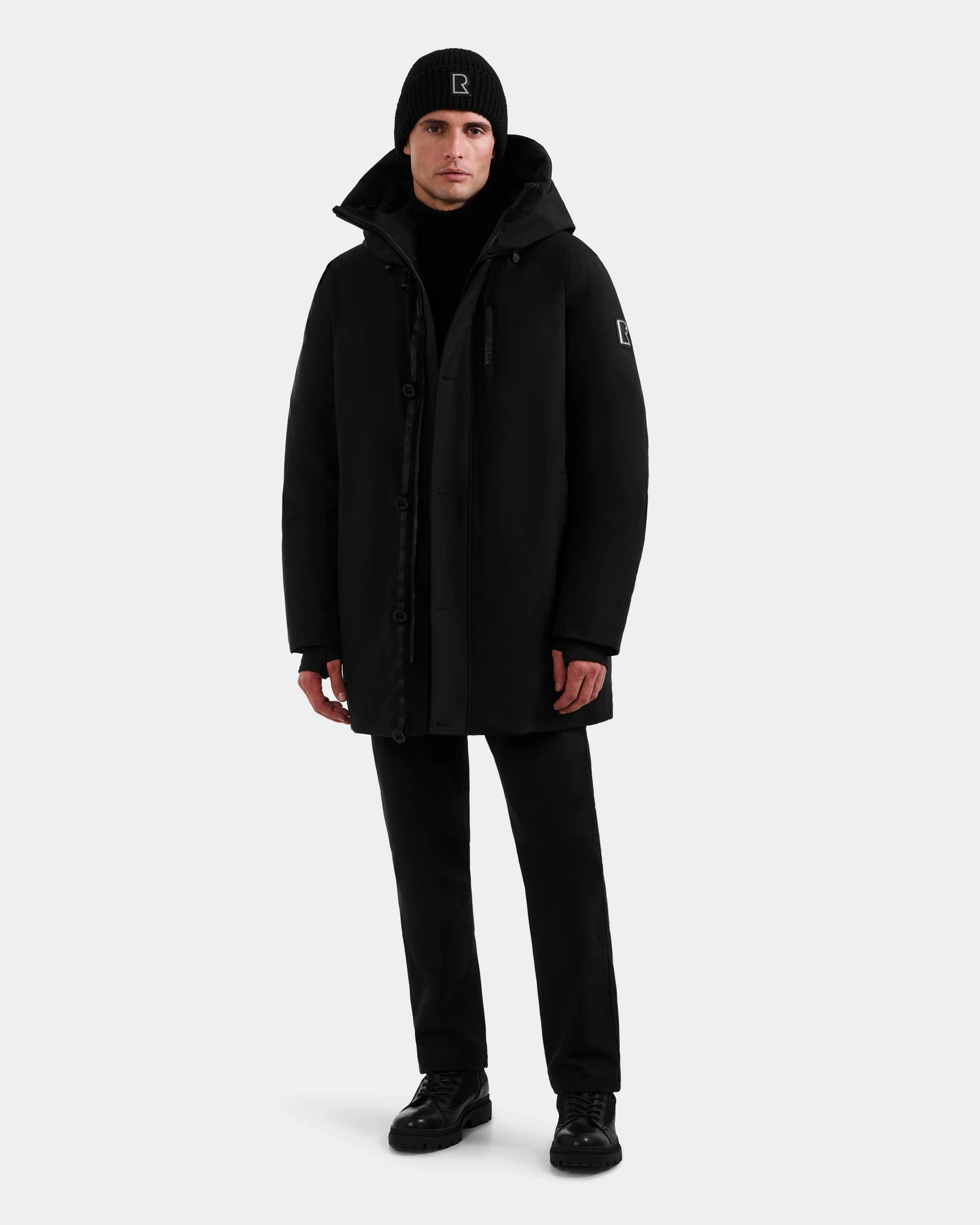 A person faces forward wearing the Rudsak COREY - M125512 Black men’s down parka, paired with black pants, shoes, and a beanie. The plain light background highlights the outfit’s sleek, warm look—ideal for cold weather.