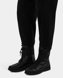 A person stands against a plain white background wearing black pants and Rudsak DAMIEN - M222500 Black men’s winter boots, which feature thick soles and weather-treated leather.