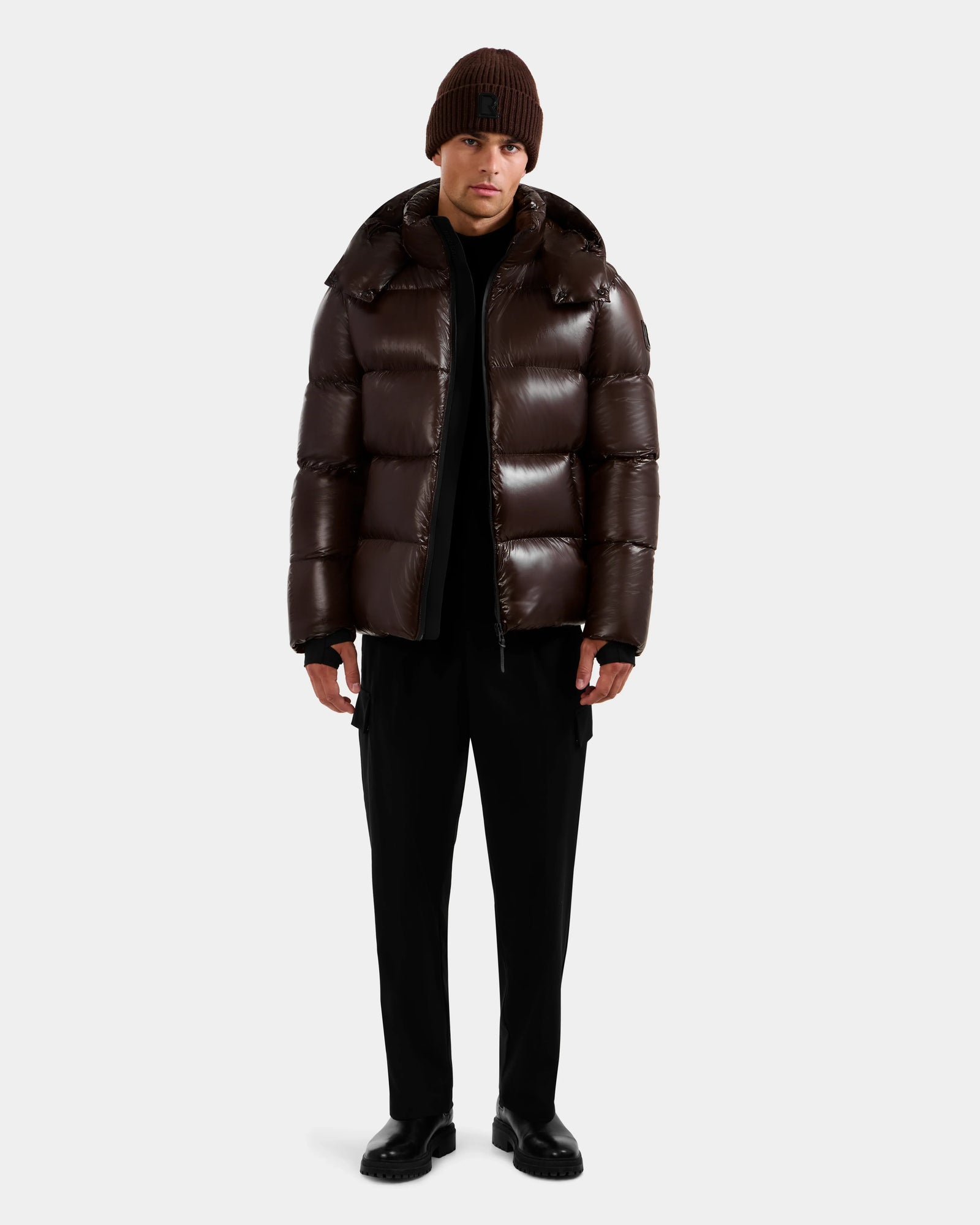 A person faces forward in a Rudsak DANIEL S - M124508 Espresso glossy, weather-resistant puffer jacket with high-loft down fill, paired with black pants, black shoes, and a brown knit beanie against a plain light background.