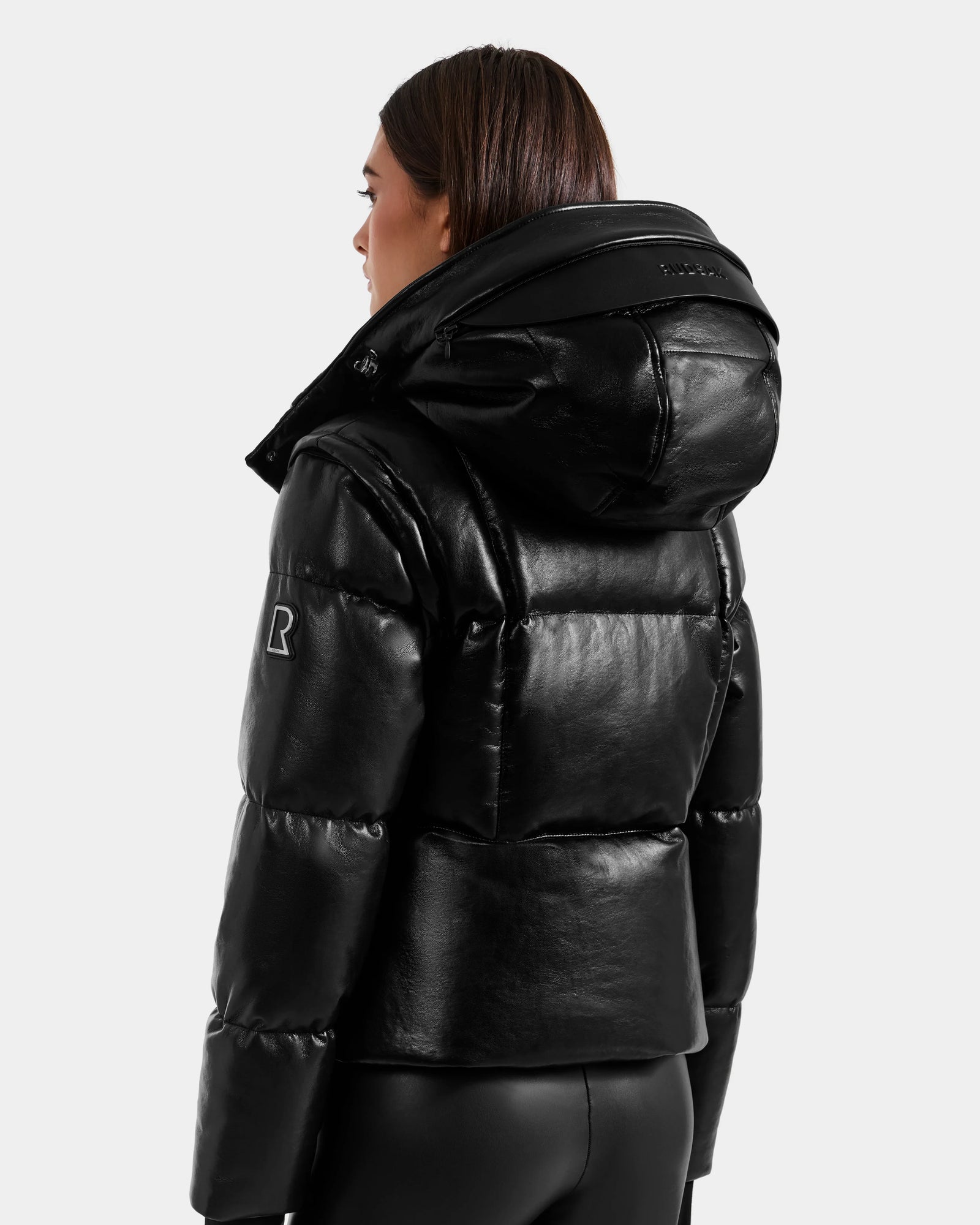 A person with straight, shoulder-length brown hair models the Rudsak DELILAH PX - L125537 Black vegan-leather puffer jacket and black pants, shown from the back and side against a plain light background.