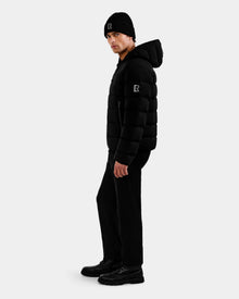 A person stands sideways in all black, wearing the Rudsak DEMIAN M - M123507 down puffer and a knit beanie, both with matching 