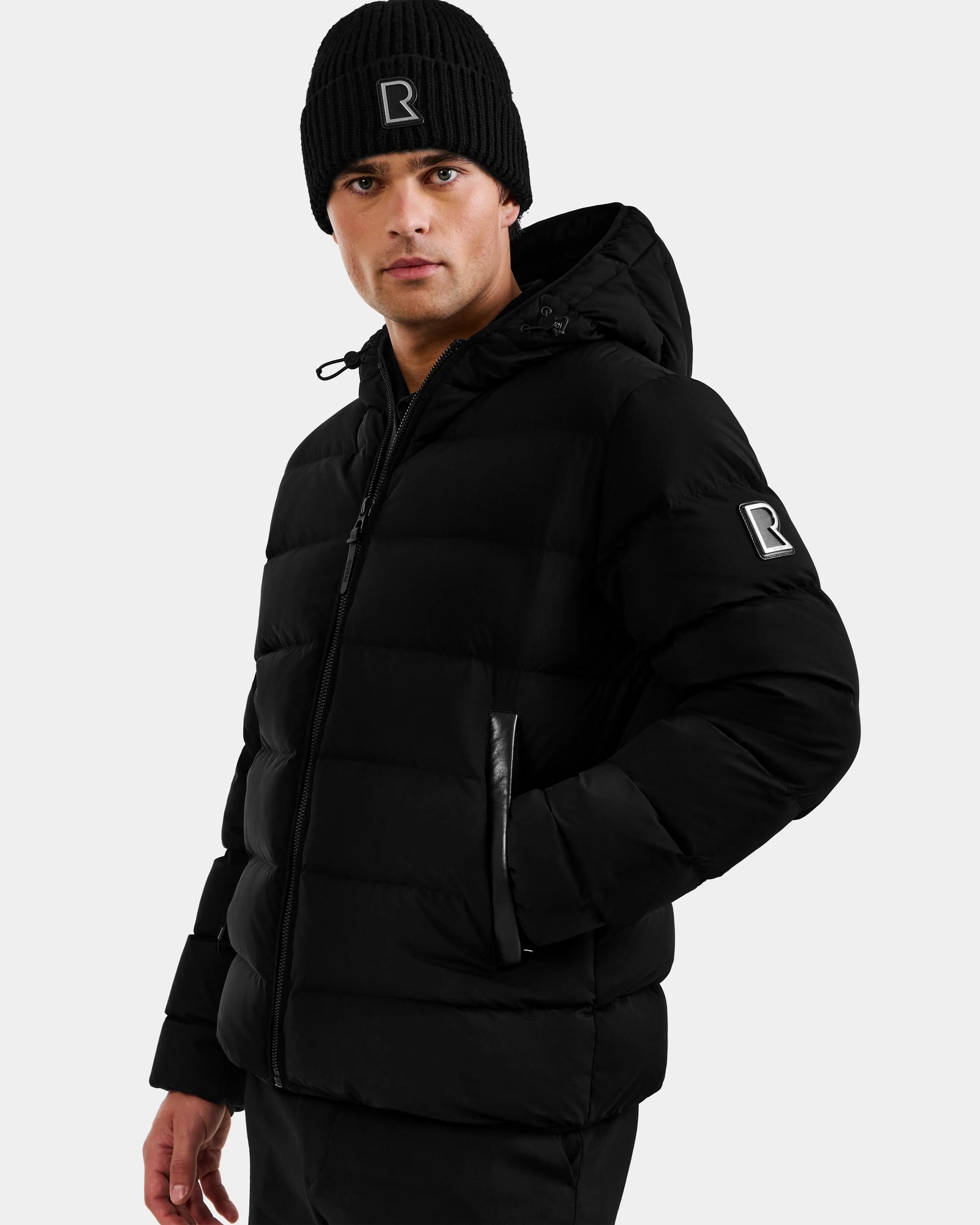A man wears the DEMIAN M - M123507 Black down puffer jacket with hood and a black knit beanie, both by Rudsak and featuring the R logo. Made from sustainable recycled fabric, he stands against a light gray background facing forward.