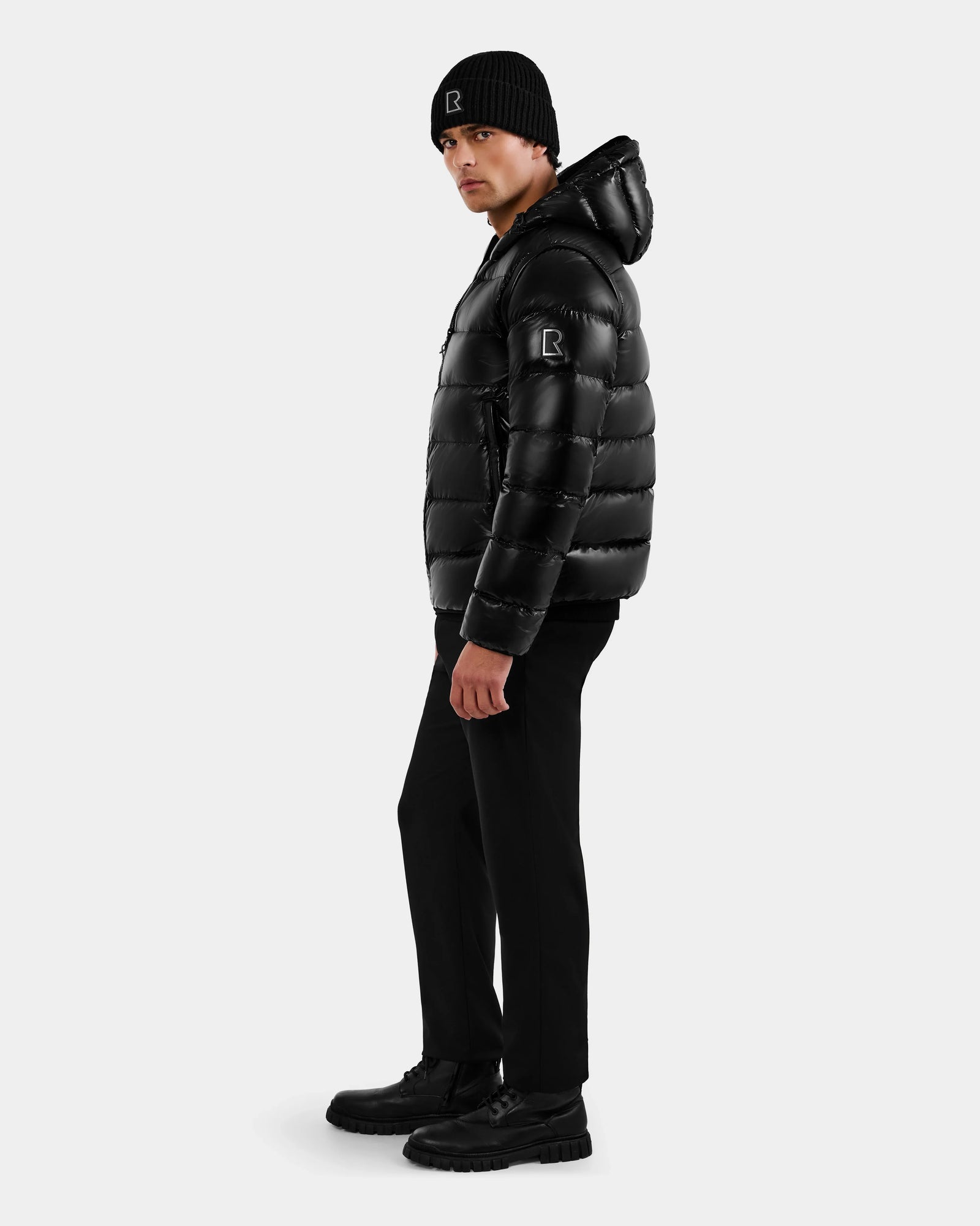 A man in the Rudsak DEMIAN S-R - M123505 Black down puffer jacket, black beanie, pants, and boots stands sideways against a plain light background, looking at the camera.
