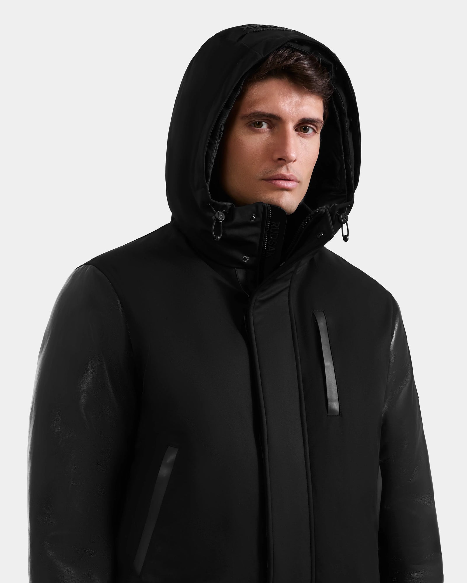A man wears the Rudsak DERON - M125539 Black jacket, featuring vegan-leather sleeves, a high collar, and zippered chest and side pockets, standing against a plain light background.