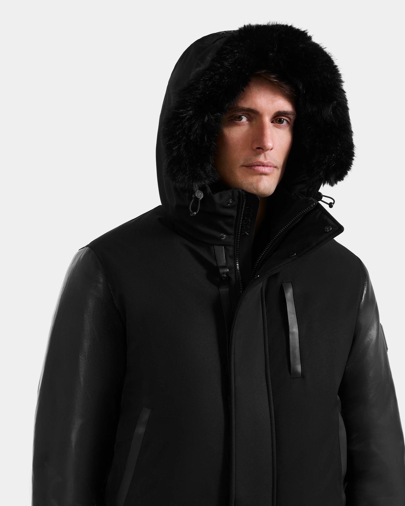 A man wearing the Rudsak DERON - M125539 Black men’s down bomber jacket with vegan-leather sleeves and a fur-lined hood stands against a plain white background.