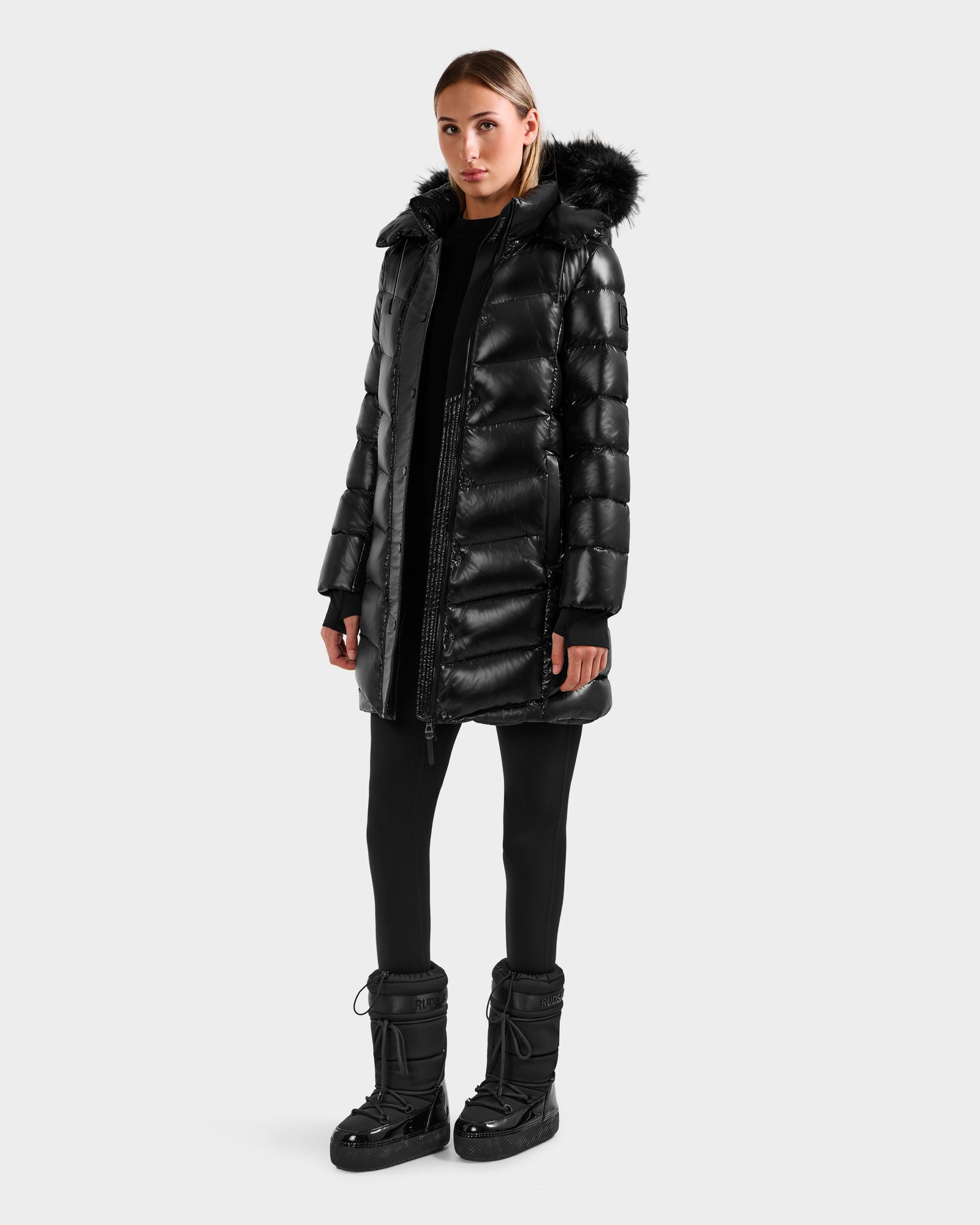 A woman stands against a plain background wearing the Rudsak ELISA S - L125572 Black quilted puffer jacket with a fur-trimmed hood, paired with black leggings and winter boots, looking straight ahead with a neutral expression.
