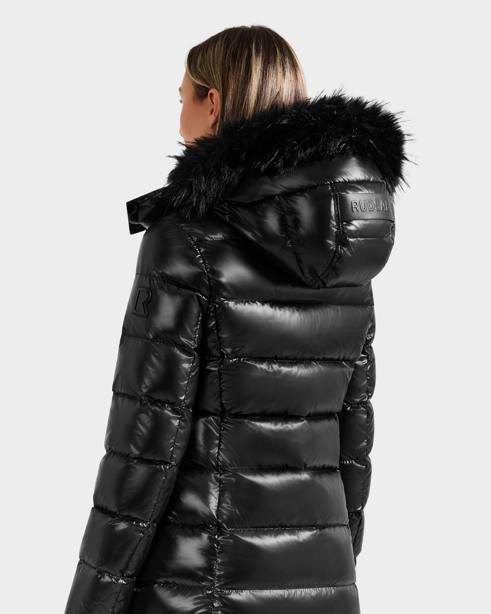 A woman is seen from behind wearing the Rudsak ELISA S - L125572 Black puffer jacket, a shiny black mid-length coat with a faux fur-trimmed hood, branded text on the back of the hood, and a patch on the sleeve.