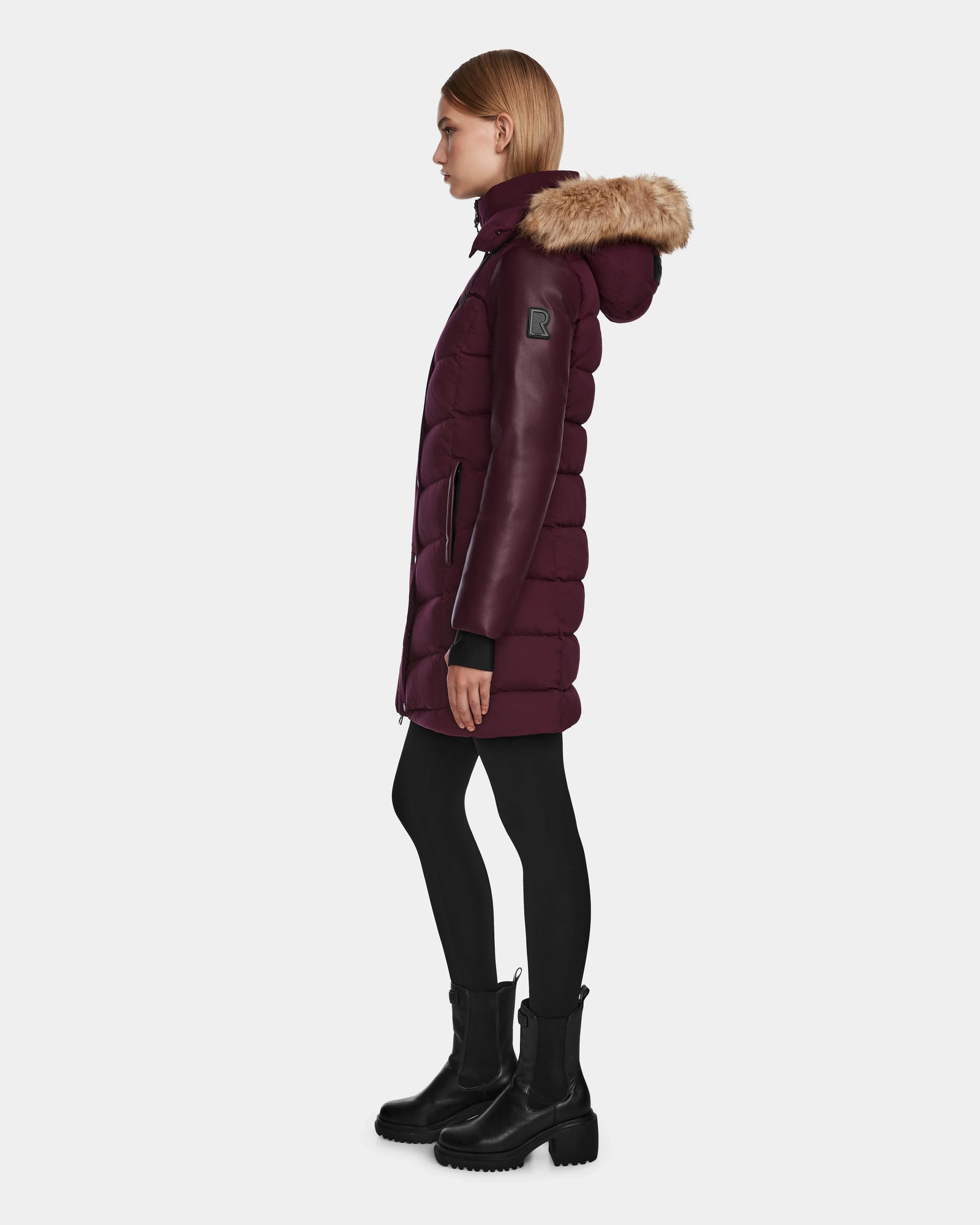 A woman stands in profile against a plain white background, wearing the Rudsak ELSA - L123535 Wine quilted down puffer with fur-trimmed hood, paired with black leggings and chunky black boots.