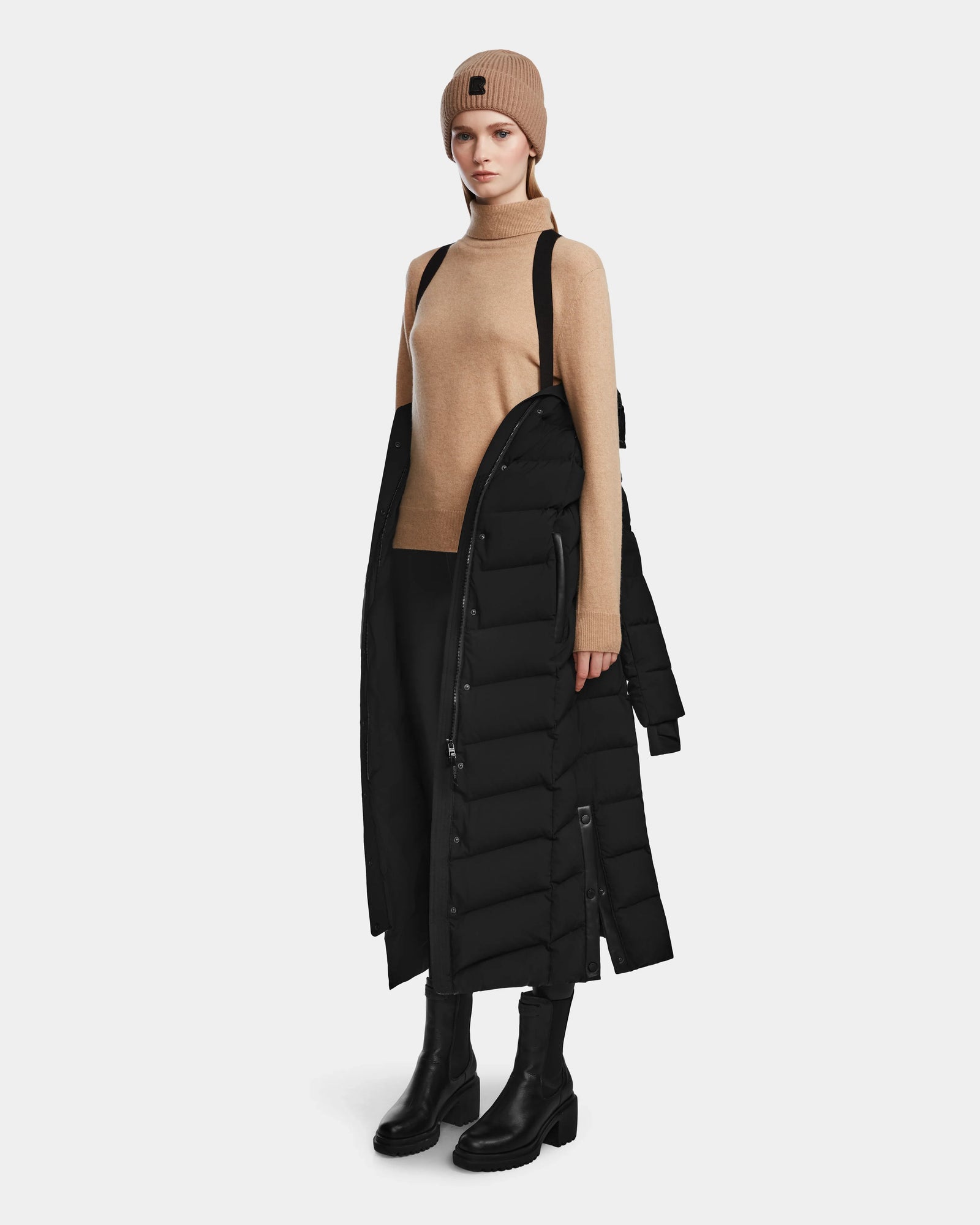A person stands against a plain white background wearing Rudsak's ELSIE M - L123517 Black/Natural women’s maxi down puffer with suspenders, a tan beanie, tan turtleneck sweater, black skirt, and black boots.