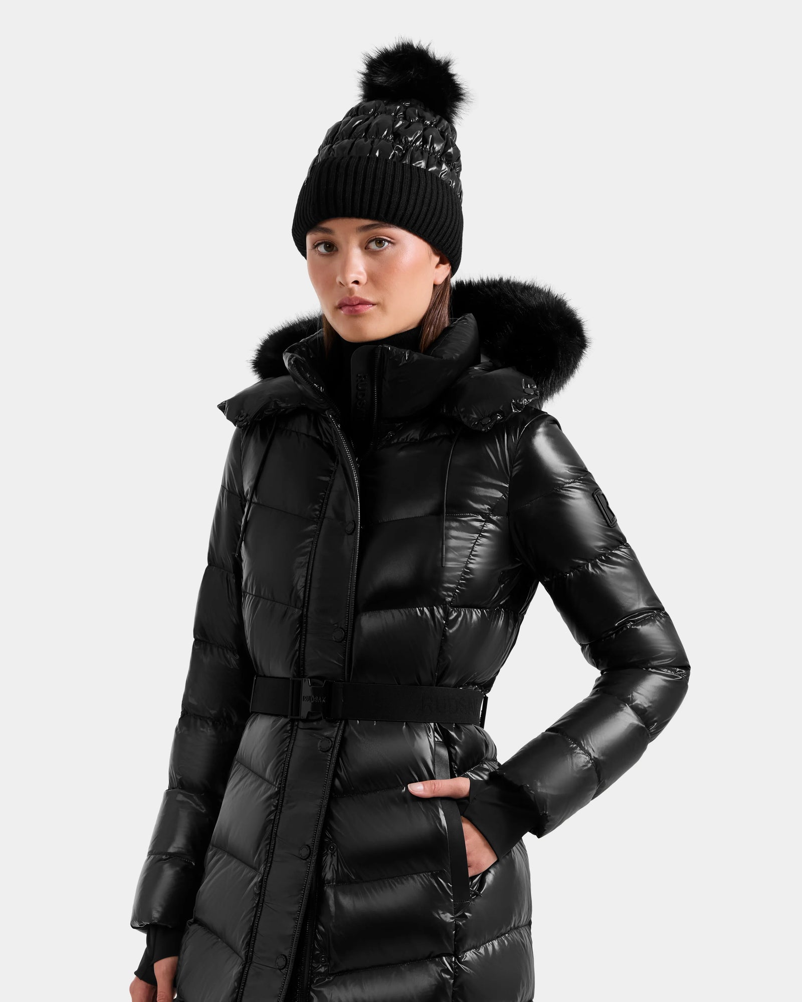 A woman in the Rudsak ELSIE S - L124536 Black water-resistant puffer coat with a fur-lined hood and a black knit pom-pom beanie, standing against a plain light background.