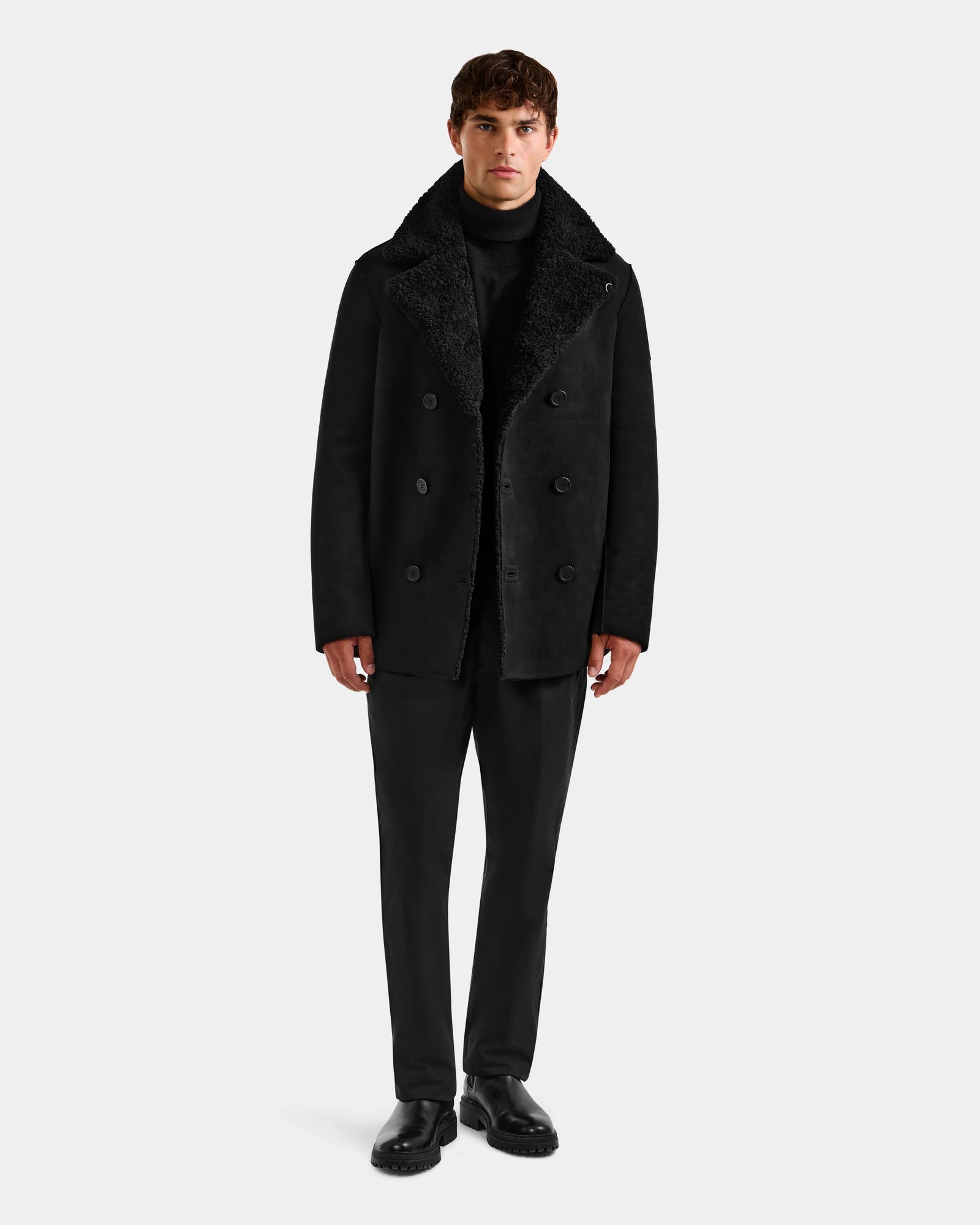 A man faces forward wearing the Rudsak ELTON - M125534 black double-breasted overcoat over a black turtleneck, trousers, and shoes against a plain light gray background.