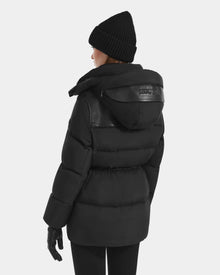 A person is shown from the back against a plain light background wearing the Rudsak EMBER M-M - L125524 Black women's down puffer with a removable hood, featuring subtle vegan-leather piping, plus a black knit beanie, gloves, and pants.