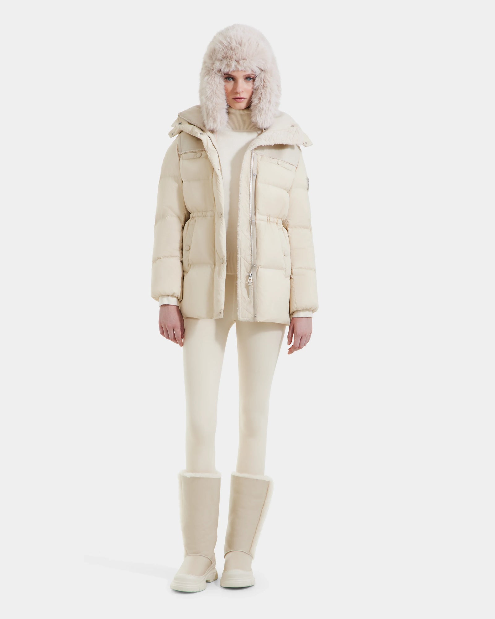 A person faces forward against a white background, wearing the Rudsak EMBER M-M - L125524 Vanilla women's down puffer in cream, matching pants, a furry hat, and tall boots.