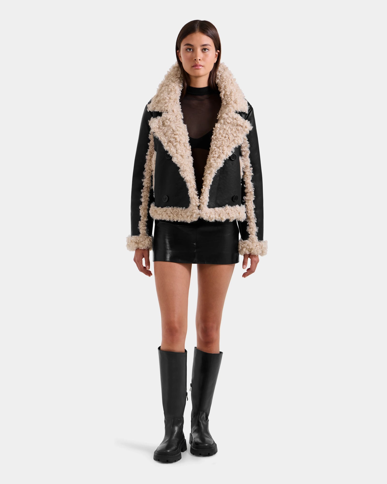 A woman stands facing forward in the Rudsak EMERY - L125548 Black vegan-leather mini skirt, a sheer black top, a women's jacket with beige faux shearling trim, and black knee-high boots against a plain light gray background.