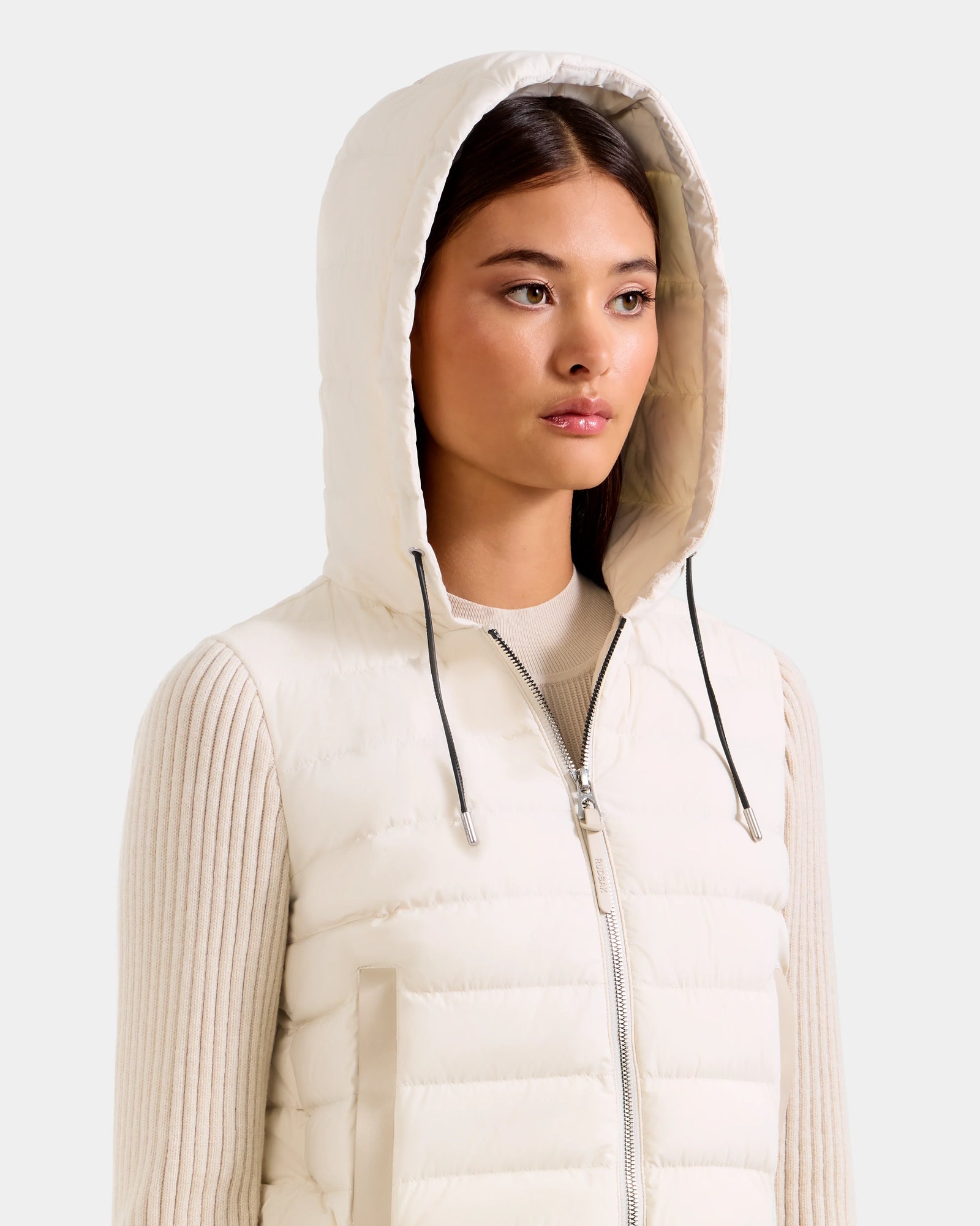 A woman wears the Rudsak EMILY - L125515 Vanilla women’s hooded down bomber over a light ribbed sweater, looking forward with a neutral expression against a plain background.