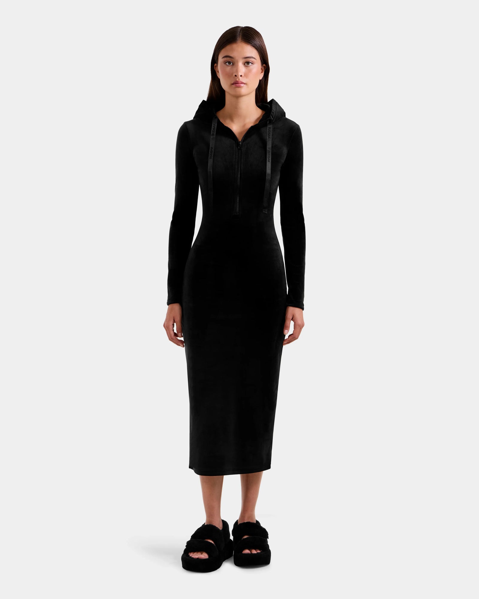 A woman faces forward in the ISABELLA - L525502 Black velour hooded dress by Rudsak, featuring a sleek midi silhouette and long sleeves, with black fuzzy slippers, set against a plain light gray background.